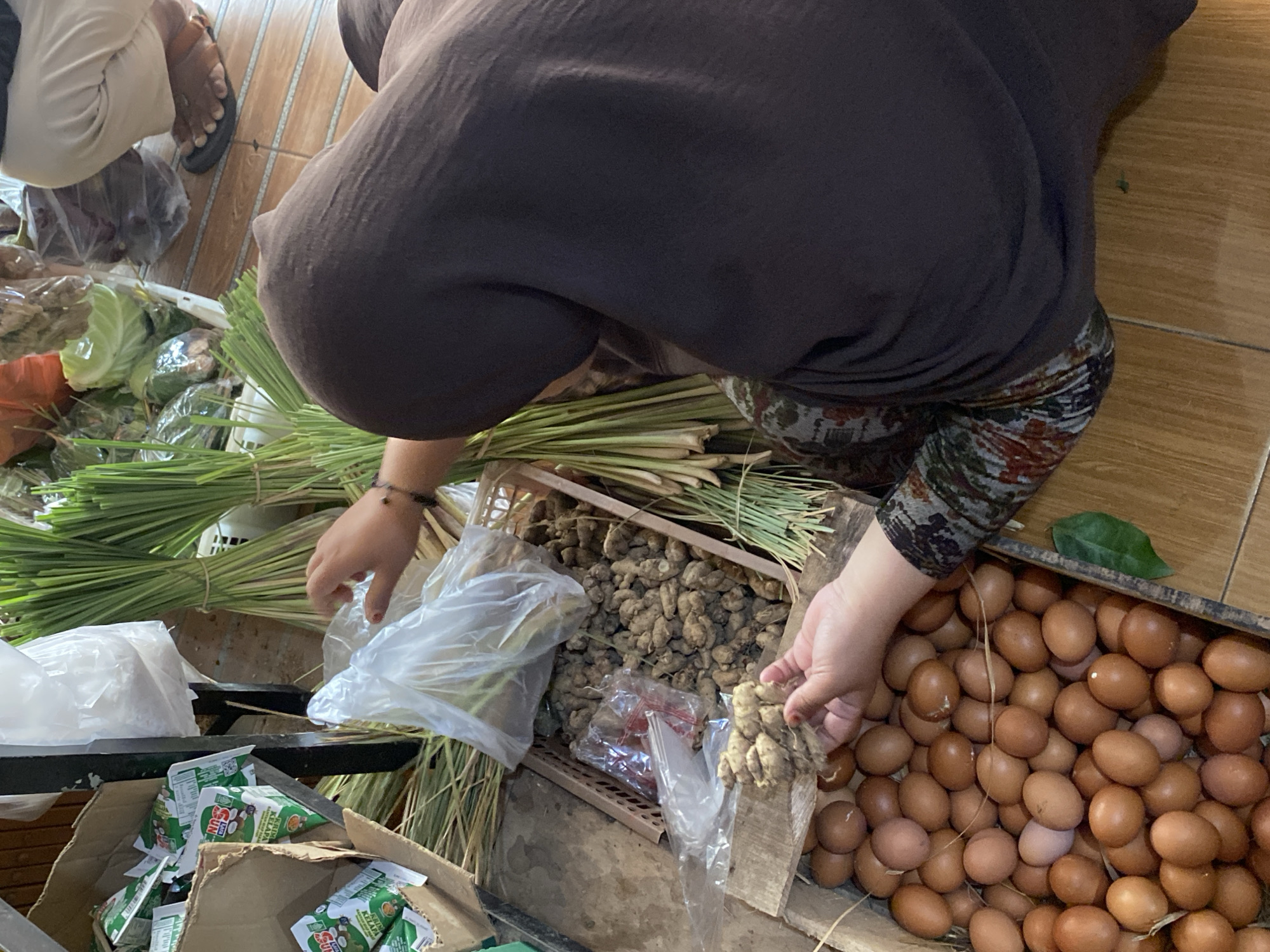 A person wearing a dark brown hijab and a patterned shirt is seen from above in a market. They are reaching into a basket of ginger. In the foreground, a crate is filled with brown eggs. To the left, there are cardboard boxes containing green and white packaged goods. Behind the person, bundles of long green stalks, likely lemongrass, are visible, along with some leafy green vegetables in clear plastic packaging. A person's feet wearing sandals are partially visible in the upper right corner. The scene appears to be an outdoor market stall in Kasihan, Indonesia, with a tiled floor.