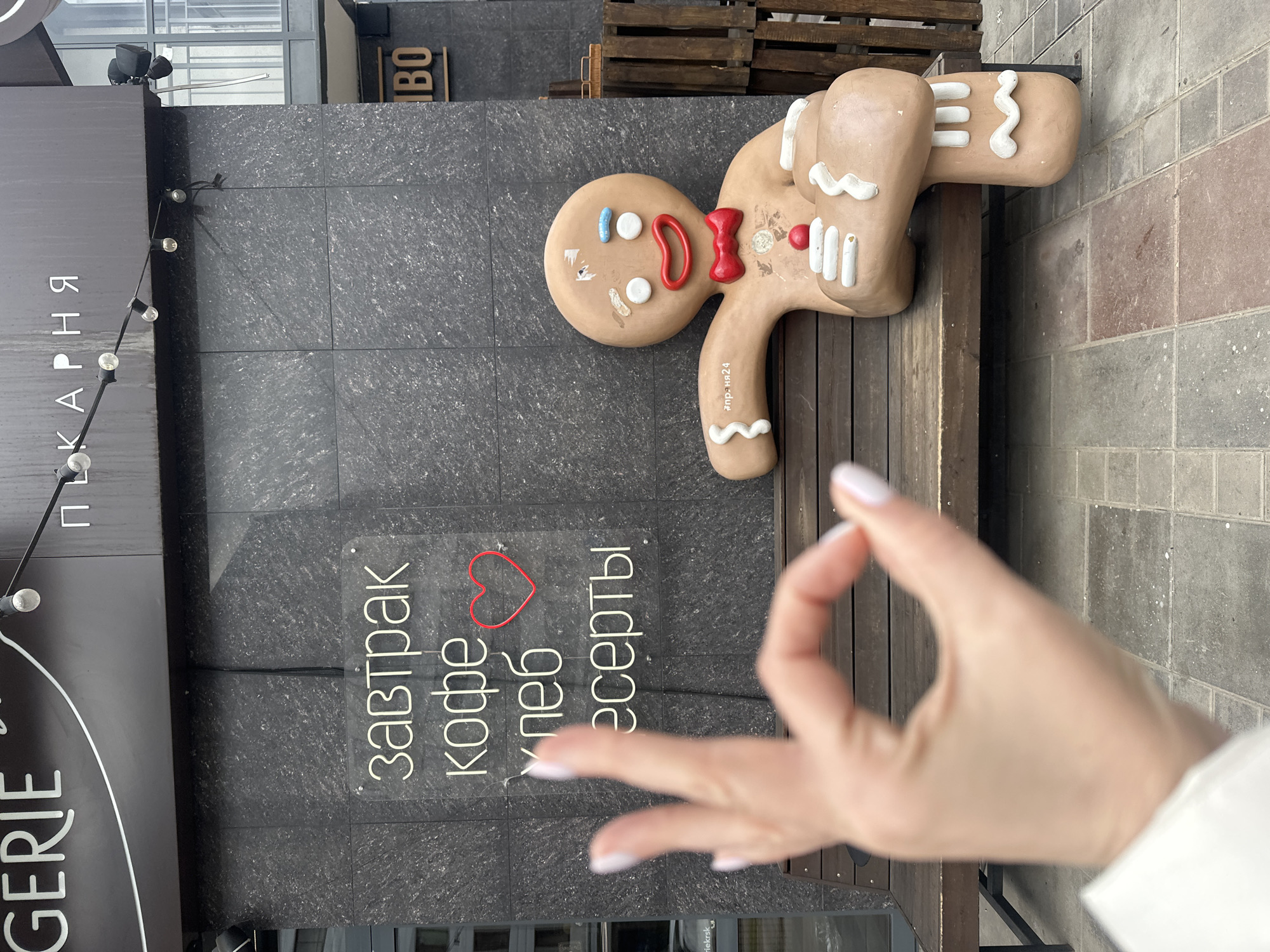The image depicts a street scene in Krasnoyarsk, Russia. A large, gingerbread-man statue rests on a wooden bench. Next to the statue is a sign in Russian that reads, "Breakfast, coffee, bread, desserts" with a heart image. A person's hand is in the foreground, forming an "OK" gesture. The statue is a prominent figure, and the lettering is vertically arranged, while the person's hand appears as a near-foreground detail. The environment appears to be a city street, as seen from the brick sidewalk and part of a building exterior.