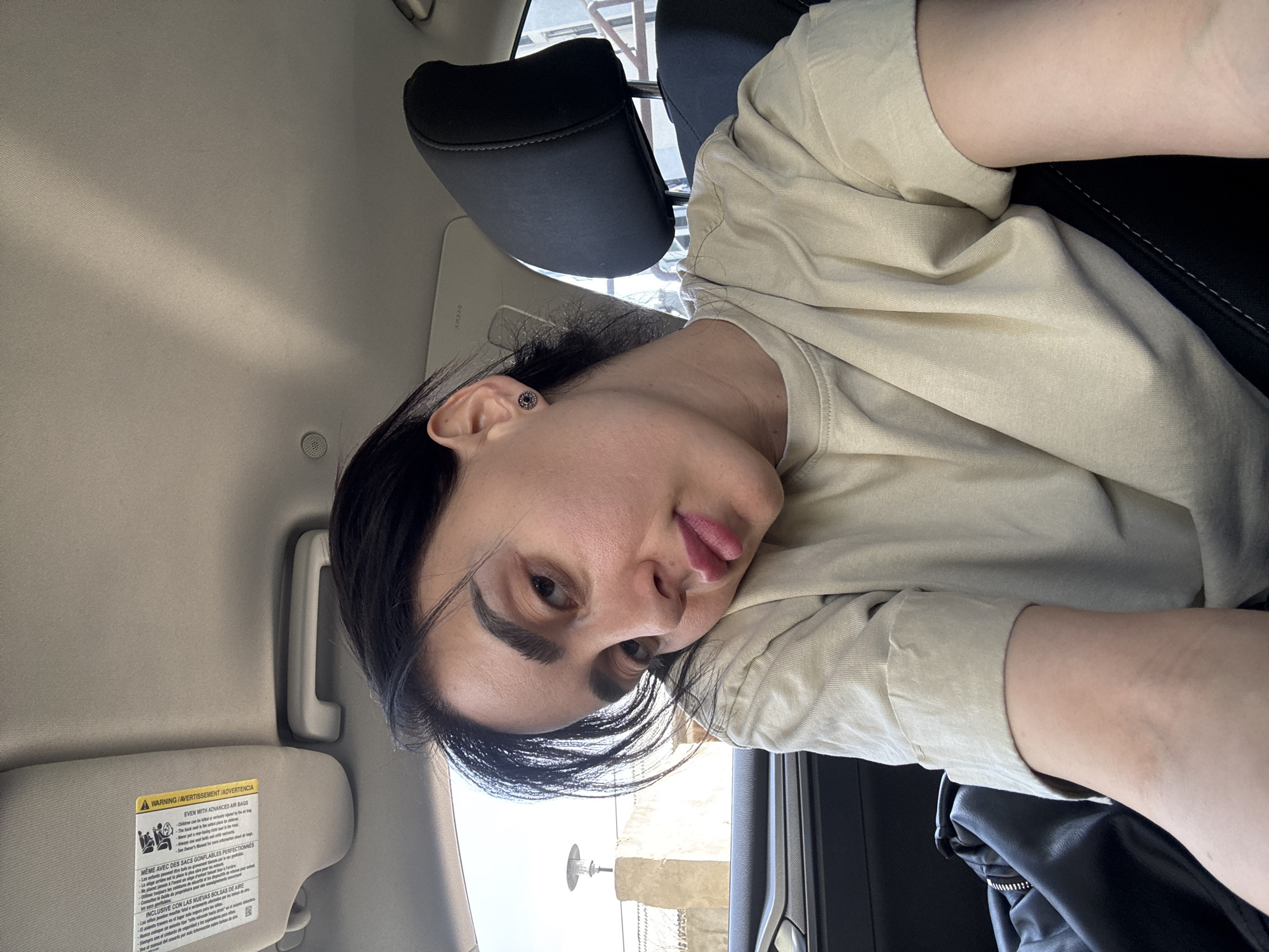 A person is sitting in the passenger seat of a car. The person has dark hair and is wearing a light-colored shirt. The interior of the car is visible, including the headrest, sun visor, and door. It's daytime and the car seems to be stationary. The location could be Tbilisi, Georgia based on a lamppost visible through the car window in the background.