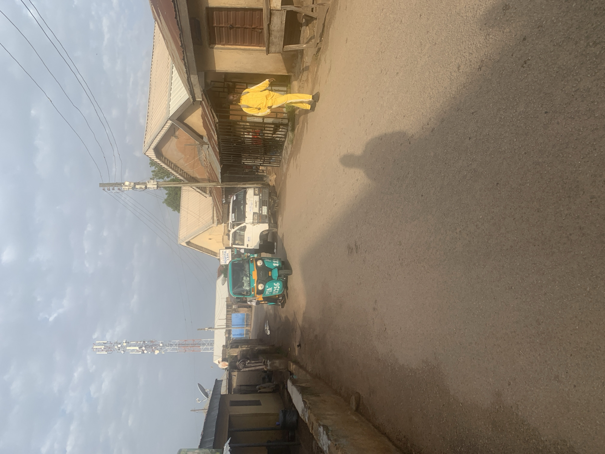 The image depicts a street scene in Jos, Nigeria, under a cloudy sky. A tall telecommunications tower is visible in the distance. The street is paved with asphalt and lined with low-rise buildings, some with corrugated iron roofs. A turquoise and green auto-rickshaw is parked on the left side of the road, displaying the license plate "PTA 11356". A white commercial van is parked behind it. On the right side of the street, a person wearing a bright yellow tracksuit and carrying a backpack walks away from the camera. The shadow of the person is cast on the road. A wooden table is set up in front of one of the buildings. The overall atmosphere is that of a quiet, possibly early morning or late afternoon scene in a modest urban area.