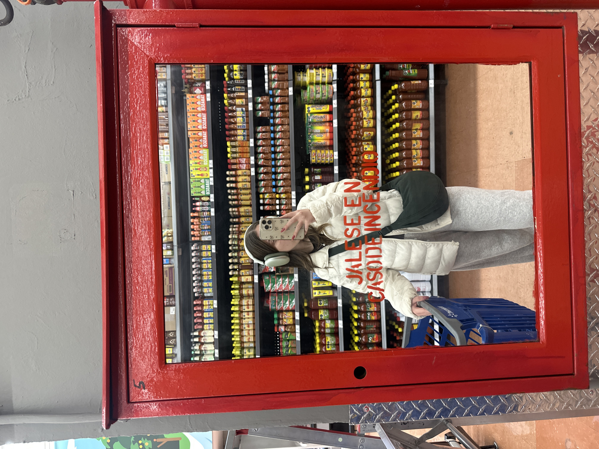 A young woman stands in front of a red-framed mirror in a store, likely in Huixquilucan de Degollado, Mexico, judging by the "JALESE EN CASO DE INCENDIO" (leave in case of fire) lettering reversed on the mirror. She is wearing a light-colored jacket, gray sweatpants, and headphones, and carrying a blue shopping cart. She holds up a phone with a patterned case to take a selfie in the mirror. The mirror reflects a display of hot sauces and other condiments. The background includes a gray wall and diamond-patterned metal.