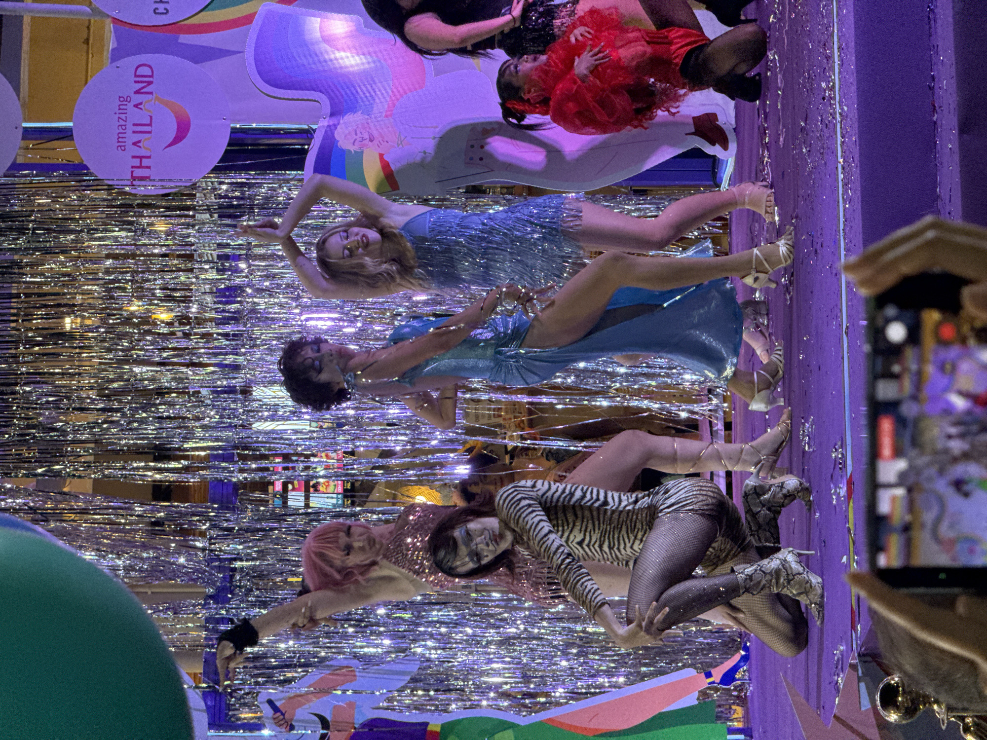 This media file depicts a vibrant and celebratory scene, likely a performance or parade. The subjects are several individuals dressed in eye-catching costumes, adorned with glitter and sequins. One person wears a shimmering blue dress with fringe, another a sparkling pink outfit, and a third is in a tiger-striped leotard and fishnet stockings. Their poses suggest dynamic movement, possibly dancing.
The setting is illuminated by bright, colorful lights, including purple and rainbow hues, and decorated with shimmering silver streamers and confetti, creating a festive atmosphere. In the background, there are circular signs with text, one prominently displaying "amazing THAILAND" with a crescent moon graphic. There are also large, abstract shapes with colorful patterns. The overall impression is one of gaiety and excitement, suggesting an event like a pride parade or a themed festival. The presence of a person holding a phone to capture the scene indicates it is a public event and a moment being documented. The time of day is likely evening or night due to the artificial lighting.