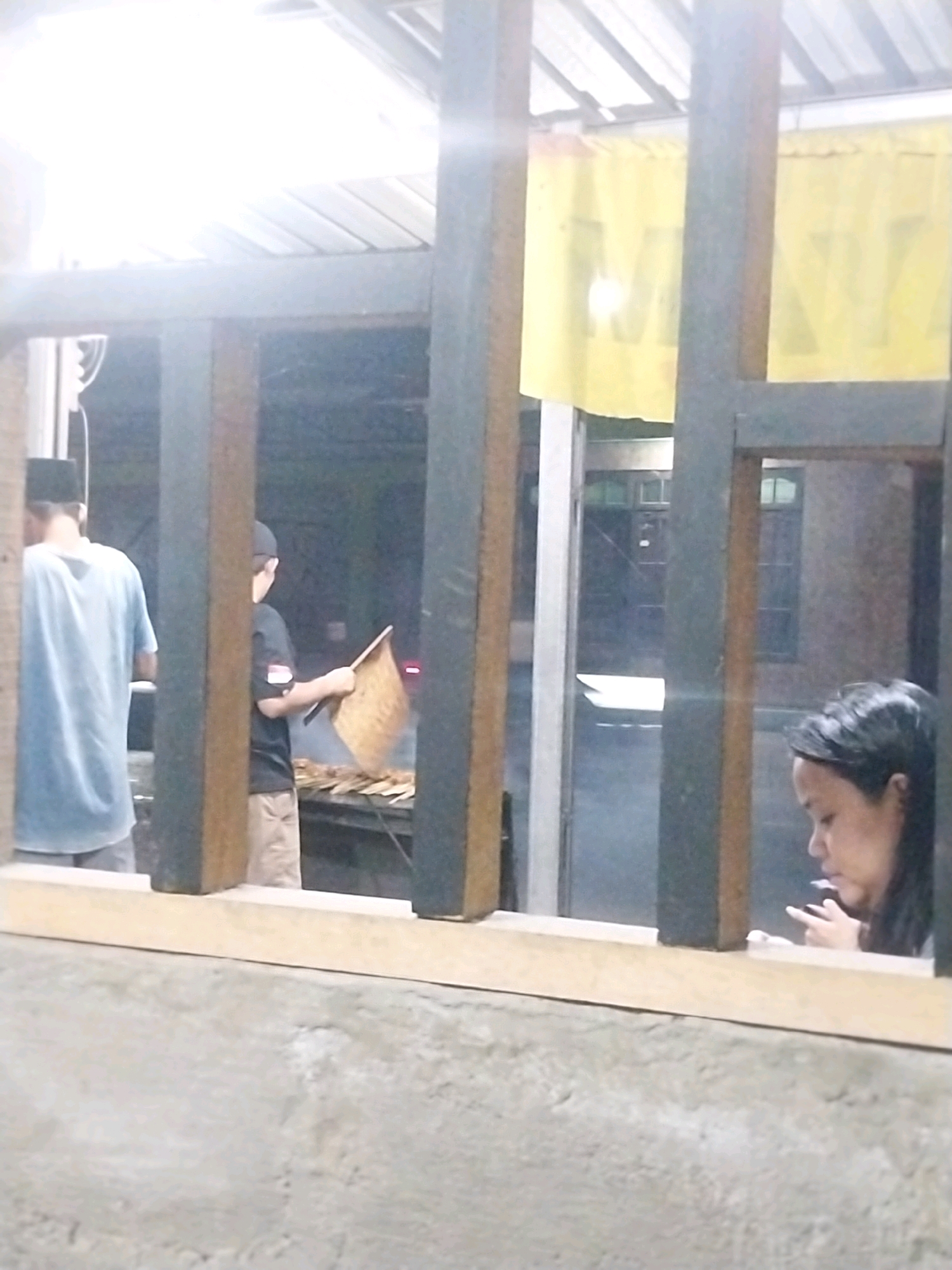 In the city of Trihanggo, Indonesia, this image captures a scene through a rustic, weathered wooden window frame. Inside, two individuals are visible. To the left, a person wearing a blue t-shirt and a black cap is seen from the back. In the center, a boy in a black t-shirt and a dark cap is holding a woven basket and appears to be interacting with a rack of skewers, suggesting a food preparation area. In the foreground on the right, a woman with dark hair is looking down, holding what appears to be a cigarette or a small object in her hand. The background is dimly lit, with hints of buildings and possibly a yellow banner with indistinct lettering. The overall setting suggests an outdoor or semi-outdoor eatery or market stall.