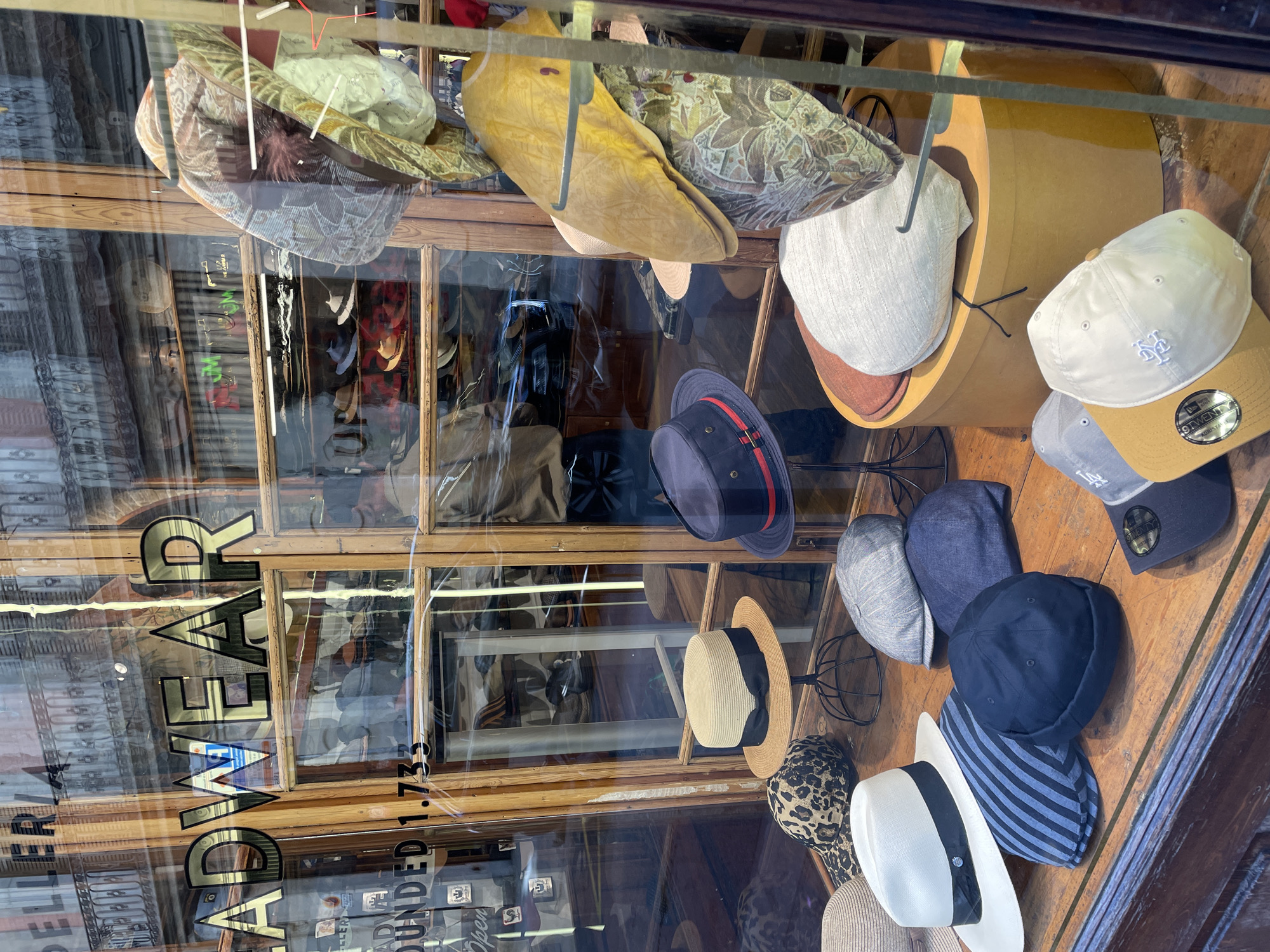 An eye-level view captures a traditional hat shop window, filled with a diverse collection of headwear. The image is rotated clockwise, showing the window vertically. Inside the wooden-framed display, various styles of hats are arranged on stands and shelves, presenting a wide selection.
Among the visible hats are several flat caps, including patterned and plain white/cream designs, as well as a yellow one. Baseball caps feature prominently, with one beige/gold cap sporting an "NY" logo and another grey cap displaying an "LA" logo. A tag on one baseball cap clearly reads "NEW ERA 9TWENTY ADJUSTABLE". Other styles include a dark blue pork pie hat with a red stripe, straw boater hats with black ribbons, leopard print and blue striped bucket hats, and a dark blue beanie or docker hat. The shop's interior, with more hats and display cases, is visible through the glass.
The activity depicted is the display of goods for sale in a shop window, intended to attract customers. The time of day appears to be daytime, suggested by the bright ambient light and the strong reflections on the glass. These reflections show parts of buildings and possibly street scenes from across the street.
Notable text includes large, vertical gold letters on the left side of the window spelling "HATWEAR". Below this, also vertically, reads "FOUNDED 1733", indicating the shop's long history. The traditional wooden window frames suggest an established business, possibly in an older district, consistent with a location like Barcelona, Spain.