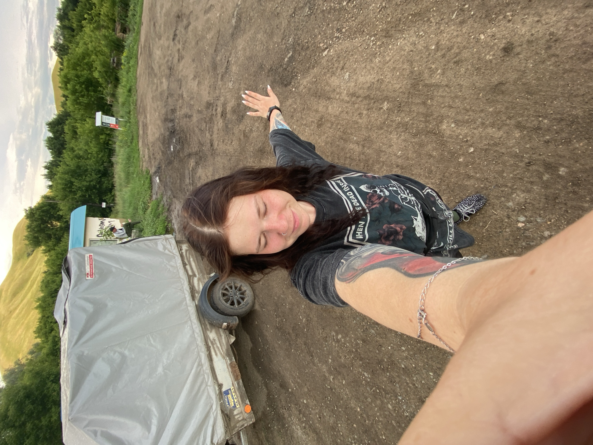 In Верхнеозёрное, Russia, a young woman with long brown hair, wearing a dark t-shirt with a graphic print and patterned sneakers, is taking a selfie outdoors on a dirt ground. She has a tattoo on her left arm and is wearing a silver bracelet. To her left is a grey-covered trailer. The background shows green trees and rolling hills under a cloudy sky. A small building with a blue roof is also visible.