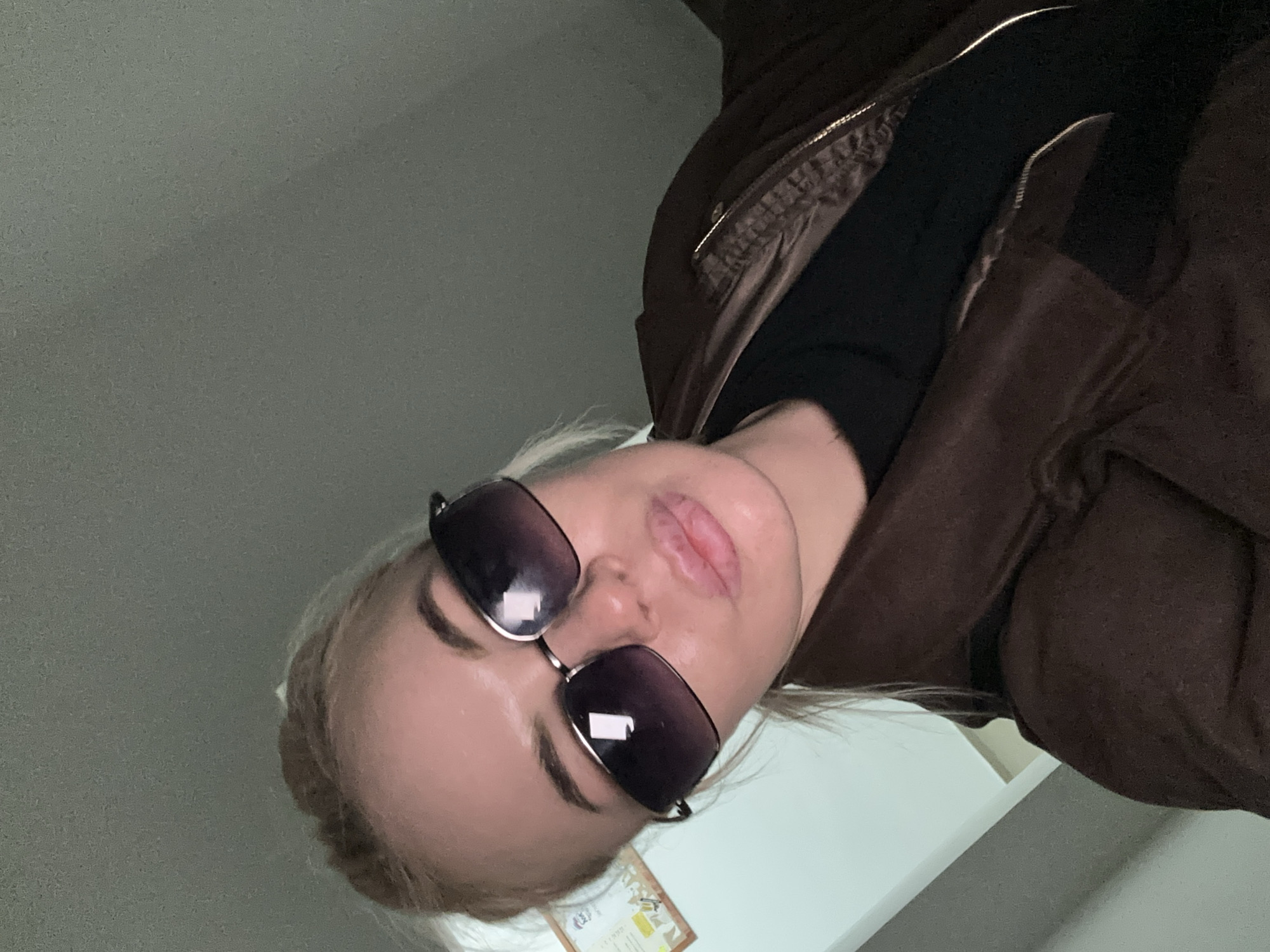 A selfie of a person with blonde hair and sunglasses, wearing a brown jacket and a black turtleneck shirt. The person is facing downward towards the camera. The location is unknown, and there is no information to suggest that the picture was taken in Высокий, Russia.
