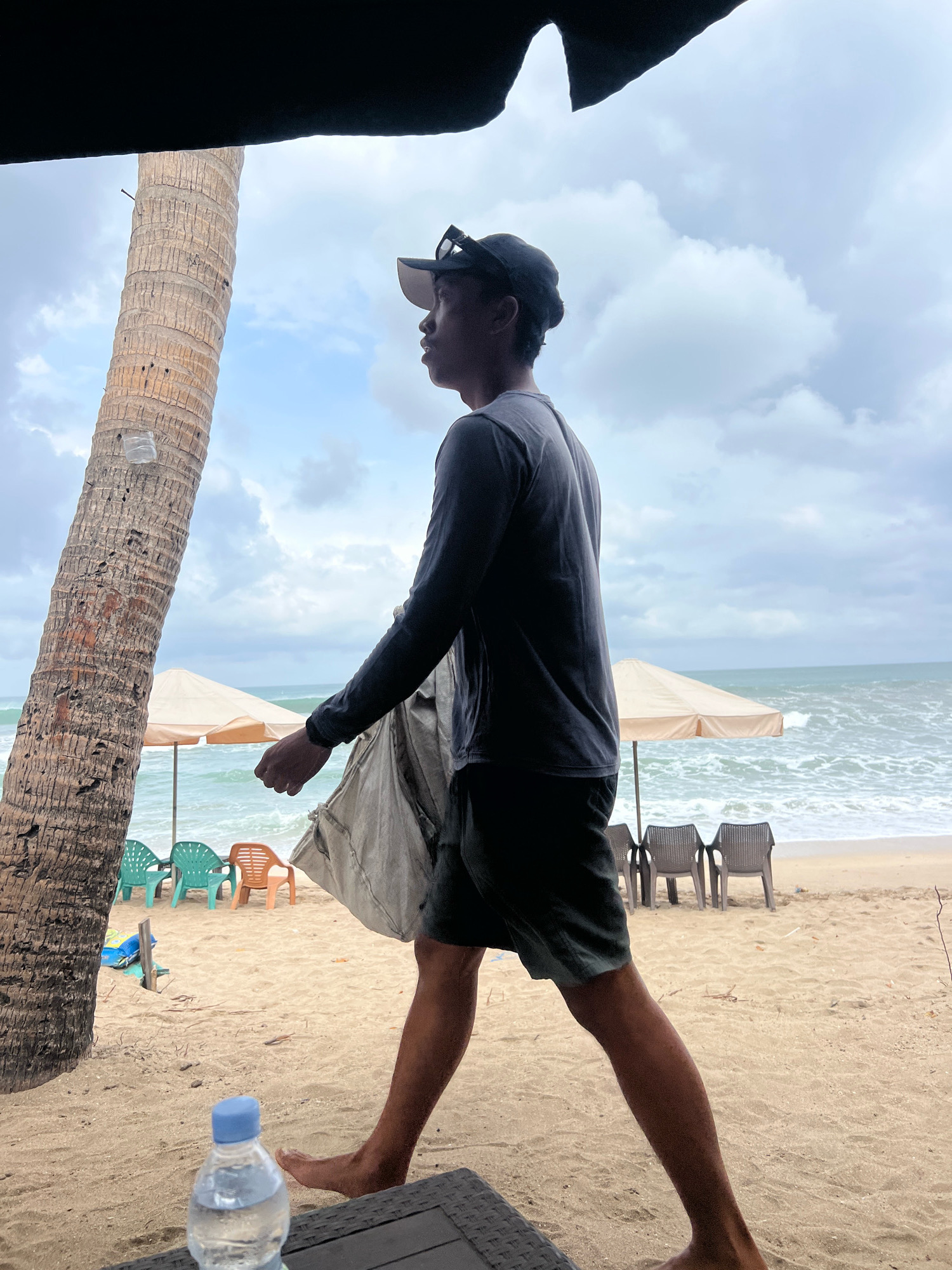 An overcast day at Kuta Beach, Indonesia, features a young man, likely in his late teens or early twenties, walking towards the ocean from a sheltered area. He is wearing a dark long-sleeved shirt and dark shorts, with a black baseball cap on his head. He is holding a light-colored tarp or bag in his right hand, with his left arm extended forward. In the foreground, a partially visible black textured surface supports a clear plastic water bottle with a blue cap. The beach sand is a light tan color, with scattered dried organic debris. Several plastic beach chairs, some green and some orange, are arranged near a beige beach umbrella. The ocean waves are moderate, with whitecaps visible. A tall, rough-barked palm tree stands to the left of the frame, with a clear plastic bottle adhered to its trunk. The overall atmosphere suggests a casual beach setting on a cloudy day.