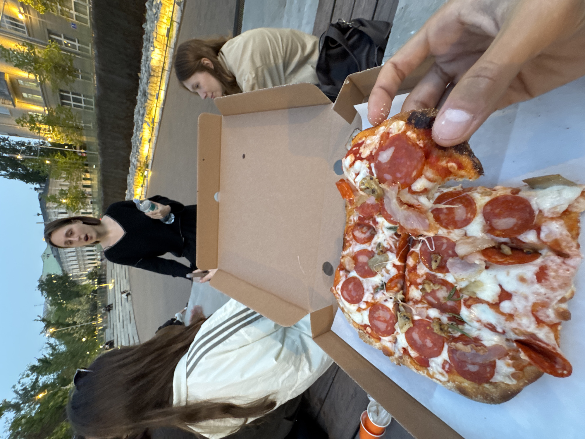 The image depicts a group of people sharing a pizza outdoors, likely in a public urban space. A pizza box sits open, revealing a pepperoni pizza with a visible hand holding a slice. Three people are partially visible around the pizza: one in a beige shirt, one in a black shirt holding a water bottle, and another in a white striped jacket. The background shows a city landscape, including buildings with lighted details. The scene is in the city of Moscow, Russia.