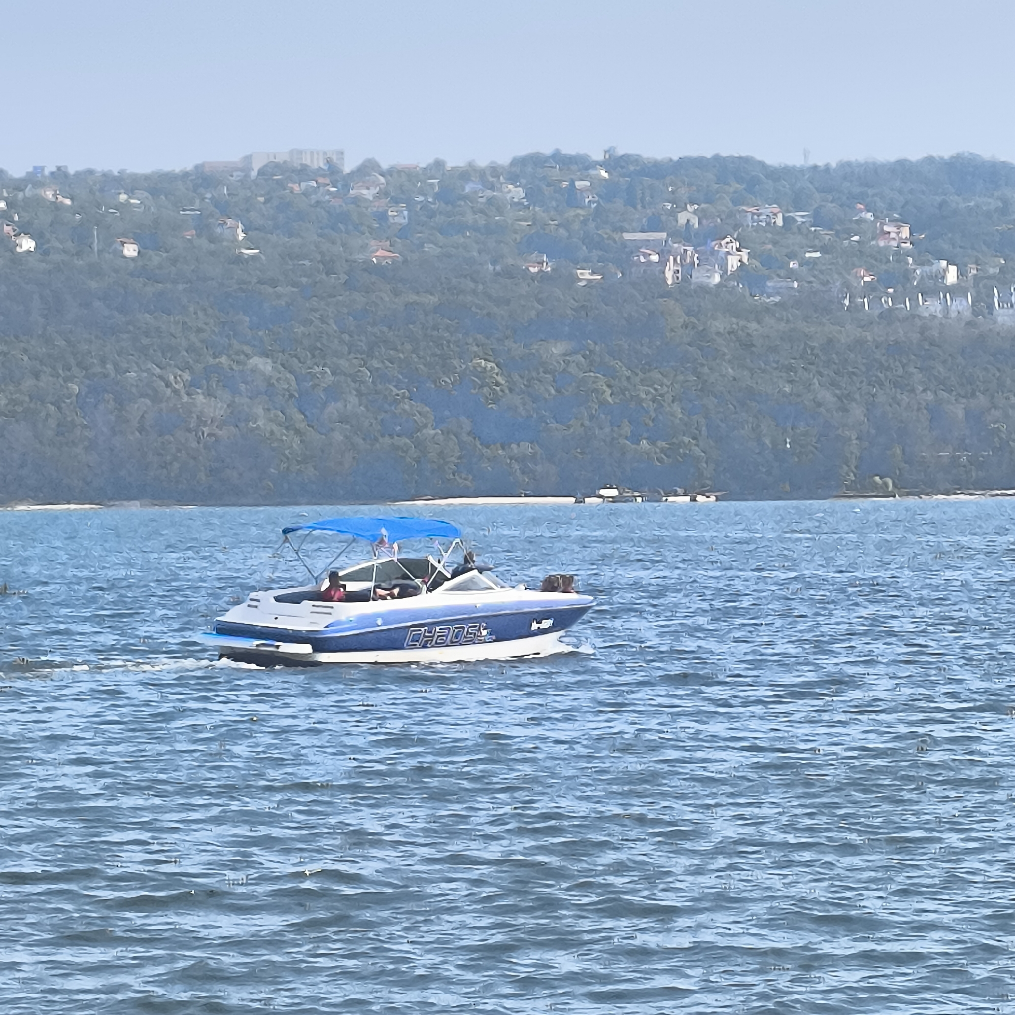 The image shows a wide, choppy body of water with a blue-canopied speed boat in the foreground, moving from left to right. The boat has a white hull with blue trim and lettering that reads "CHAOS." There are at least two individuals visible on the boat, one at the helm. In the background, a densely vegetated hill rises, dotted with houses and buildings, indicative of a coastal city. The sky is clear and light blue. This scene is consistent with the coastline of Varna, Bulgaria, specifically likely along the Varna Bay or a nearby lake, given the urban development on the hillsides and the presence of a speed boat on the water.