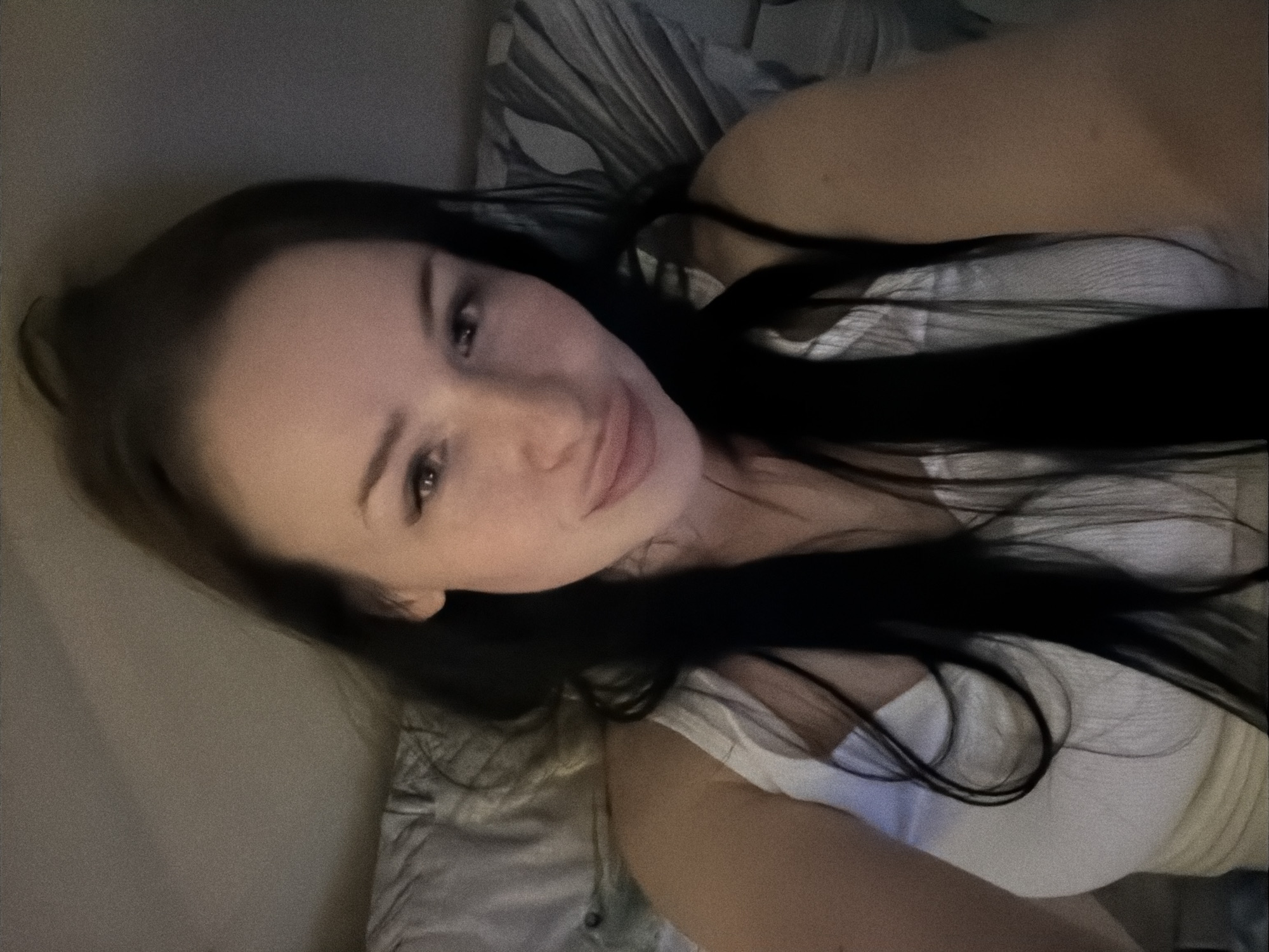 The image displays a single individual, an adult female with long, dark hair, positioned in what appears to be a bed or reclined surface. Her hair is styled partially up, with strands falling forward around her face and shoulders. She is wearing a white, ribbed tank top. Her head is tilted slightly, and her lips are pursed. Her eyes are open and directed towards the camera. The background consists of indistinct, soft-textured surfaces consistent with bedding (pillows, sheets) and a plain wall, rendered in muted gray tones. The lighting is dim, resulting in a noticeable grainy texture throughout the image. No identifying features within the image indicate a specific geographic location, including the city of Sertolovo, Russia.
