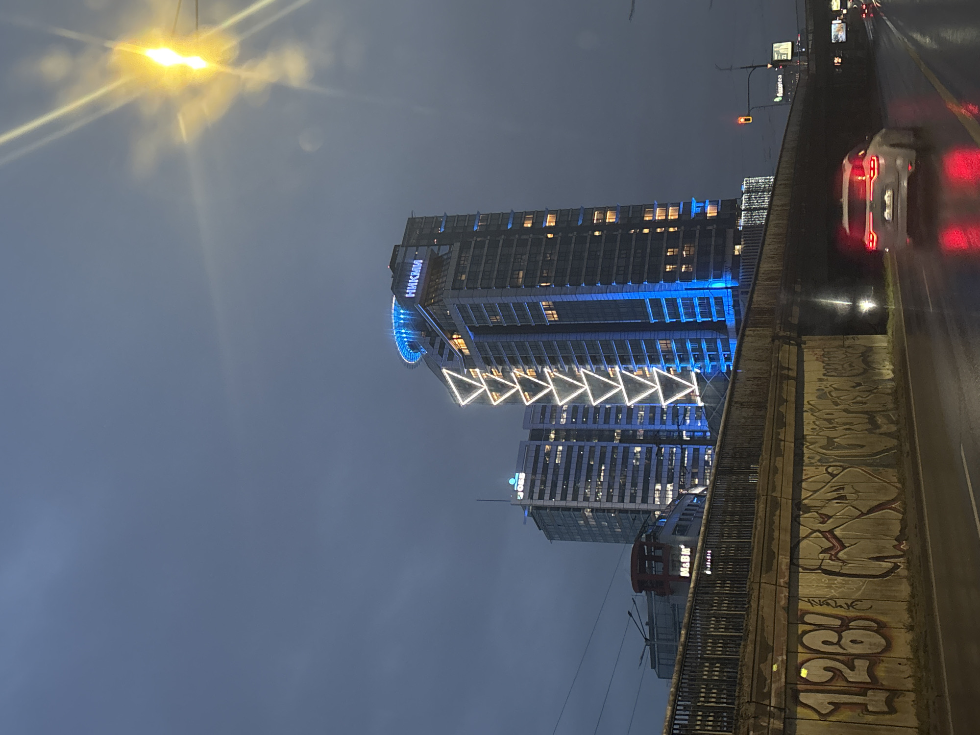 The image depicts a nighttime urban scene in Sofia, Bulgaria. The Hayatt Regency Sofia hotel is visible, illuminated with blue lights. A string of triangle-shaped lights adorns the building's facade. A road with a car is visible to the right, positioned on a bridge or overpass. The overpass is covered in graffiti. A street lamp is located to the left, emitting a bright light. The sky appears cloudy and dark.