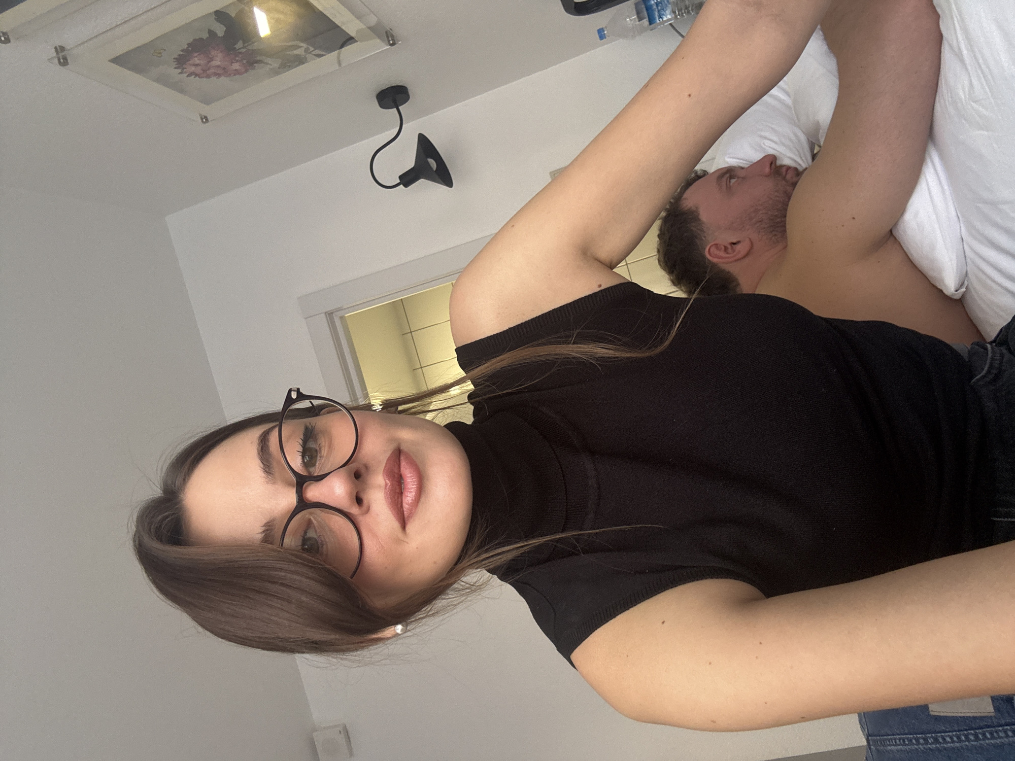 A woman wearing glasses and a black turtleneck shirt is in the foreground, seemingly taking a selfie. Behind her, a man is lying in bed with a white pillow. The room appears to be a bedroom with a white wall. A picture frame is hanging near the ceiling. A modern, black sconce is mounted on the wall. A closed door is visible in the background. The location is not discernable as Moscow, Russia, based on the visual information provided.