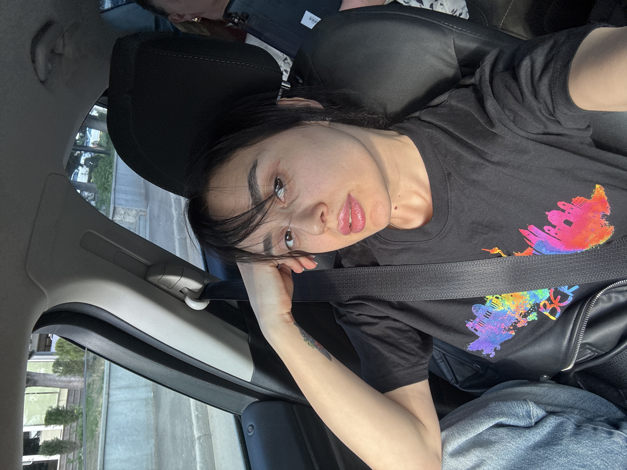 A woman sits in the passenger seat of a car, wearing a black t-shirt with a colorful city skyline design. She is secured with a black seatbelt. Her head is tilted, resting on her left hand, and she looks at the camera. Her dark hair is partially obscuring her face. The interior of the car is visible, including the seat and the car's side panel. Through the car window, a streetscape is visible, seemingly in Tbilisi, Georgia, based on architectural styles and vegetation that seem to be reflecting in the glass. A tattoo is partially visible on her left forearm. She is wearing a black jacket and blue jeans.