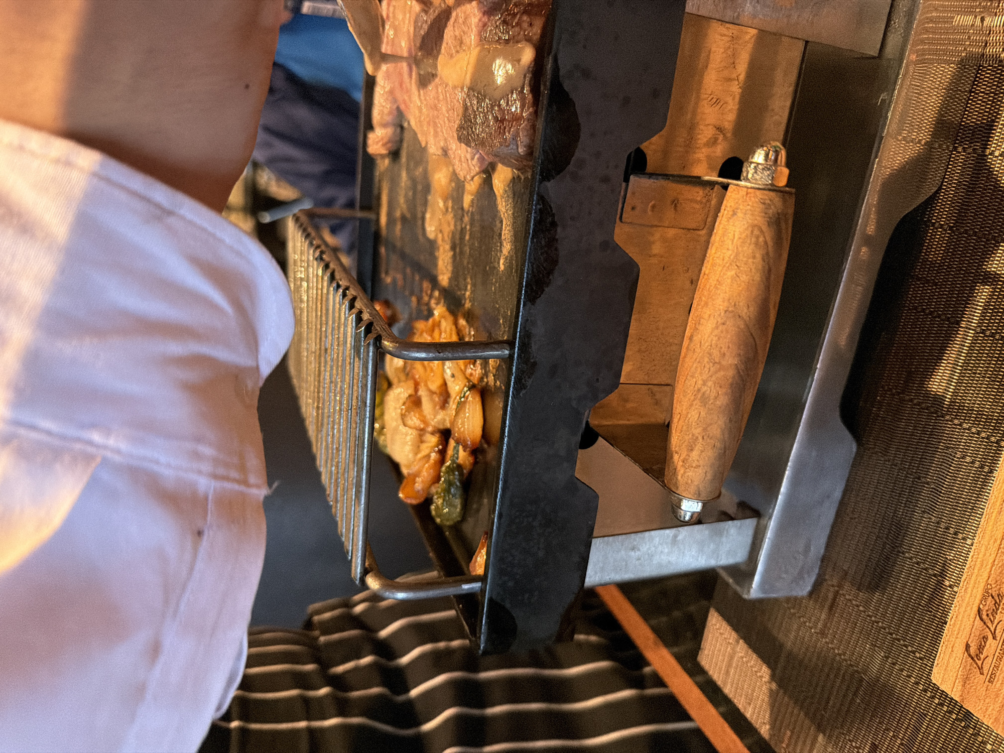 The image depicts a close-up, overhead view of a tabletop grilling experience in a restaurant located in San Bernardino Tlaxcalancingo, Mexico. In the foreground, a portable metallic grill unit with a prominent wooden handle at its front is centered on a dark, textured placemat. The unit consists of a flat, black cast-iron griddle on which food is actively cooking, and a raised, empty metal grate above it.
On the griddle's left side, various chopped vegetables, including green bell peppers and sliced onions, are sizzling in oil. On the right side, several cuts of reddish-pink meat, consistent with steak slices, are cooking. A metallic utensil, possibly a spoon or small ladle, is visible above the meat, from which a light-colored liquid or sauce is being poured or dripped onto the steak.
A person wearing a light pink or peach-colored top is partially visible in the upper right quadrant, with their right arm and shoulder prominent above the grill, suggesting active participation in the cooking process. To the far left, a person wearing a black and white striped apron is partially visible, indicating a server or chef. In the blurred background, another individual's arm and shoulder, clad in a blue garment, are faintly discernible.
In the bottom-left corner of the image, partially obscured, is a small wooden card or plaque with text identifying "Lomo Lindo GRUPO" and "RESTAURANTE," confirming the commercial dining setting. The scene is illuminated by warm, artificial lighting, casting reflections on the griddle surface and the cooking food.