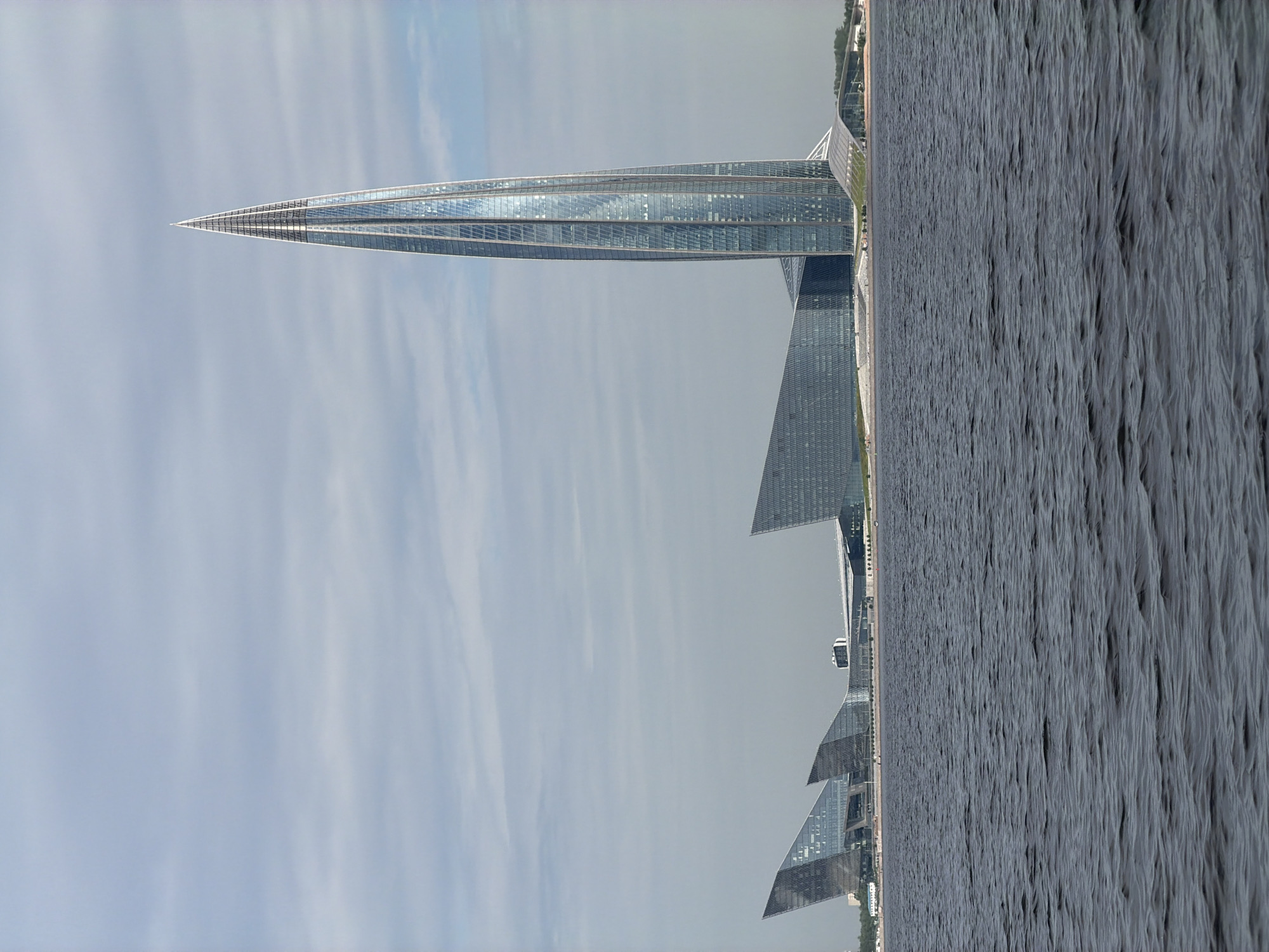 A vertical, medium shot captures a cluster of modern, glass-faced buildings situated on the waterfront of Saint Petersburg, Russia. In the foreground, the dark, choppy surface of a body of water, likely the Neva River or the Gulf of Finland, occupies the lower half of the frame. The water is a deep gray, with subtle ripples and waves indicating a slight breeze.
Dominating the right side of the image is a very tall, slender skyscraper with a distinct, pointed spire. Its facade is composed of glass panels, reflecting the sky and surrounding architecture. The skyscraper tapers to a needle-like point at its apex.
To the left of the skyscraper, a series of large, angular buildings with sloped, glass facades are visible. These structures have a striking, geometric design, resembling triangular or trapezoidal shapes that rise from the waterfront. One of these buildings appears to have multiple tiered sections. Further to the left, additional buildings form a low-rise complex that stretches along the embankment.
The buildings are set against a backdrop of a pale blue sky filled with scattered, wispy clouds. The overall impression is one of modern architectural development within the historical context of Saint Petersburg. There are no people visible in the image.