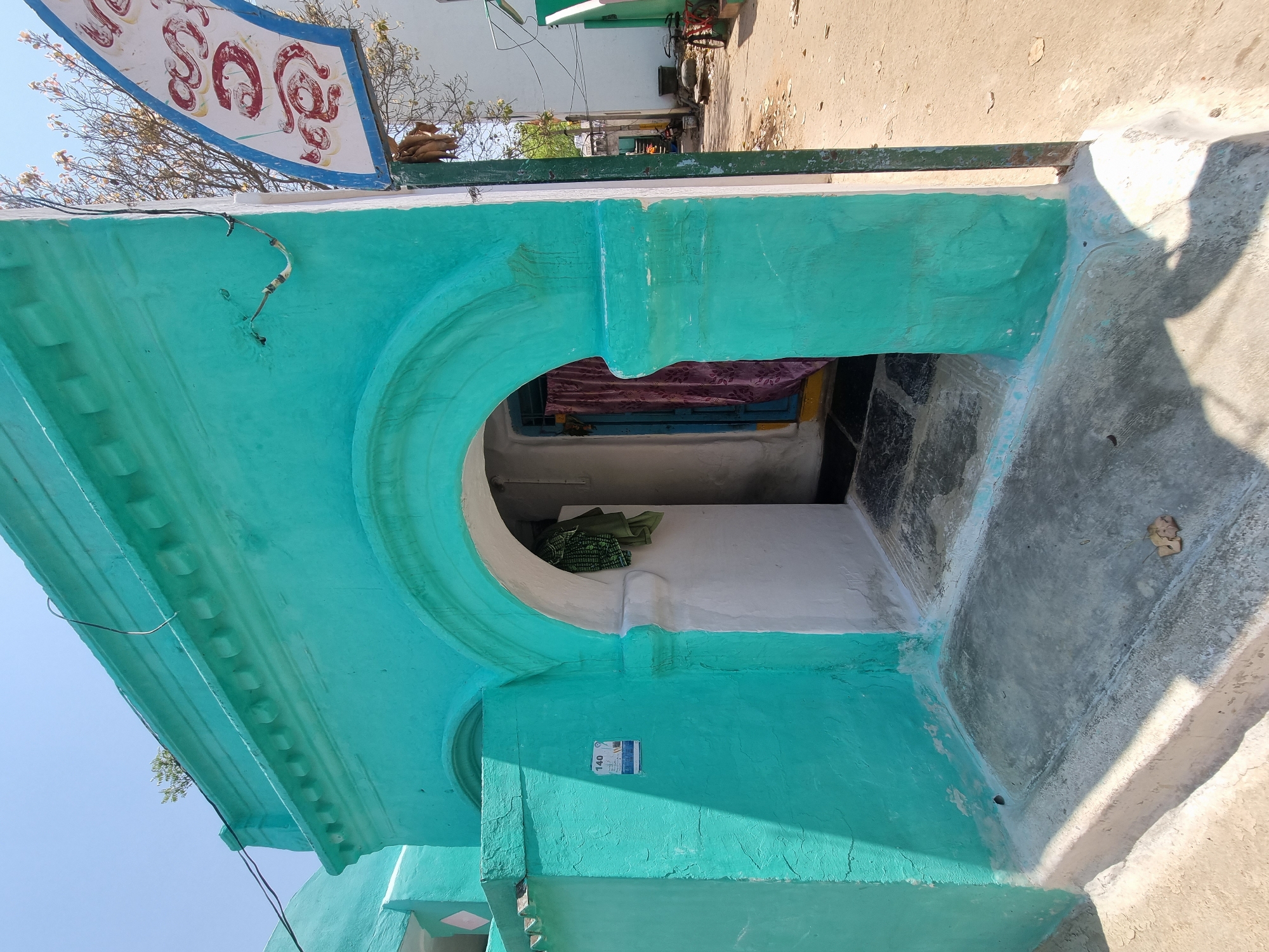 An outdoor scene in Pedda Kammavaripalli, India, displays the facade of a building painted predominantly in a weathered turquoise hue under a clear blue sky. The central feature is an arched entrance, painted turquoise on the exterior and white within the archway. Inside the arch, a dark blue door is partially obscured by a purple curtain. A green garment hangs on the white wall inside the entrance. The floor within the archway is dark tiled.
To the left of the arch, a turquoise wall section features a small white rectangular sign with the number "140" and additional faded text and graphics. Above the arch, the building's parapet is adorned with horizontal moldings and decorative rounded protrusions. A small green plant grows atop this parapet. Electrical wiring, dark in color, extends across the upper facade and descends along the right side.
The ground directly in front of the entrance consists of a concrete slab, with steps leading down to a dirt path. Strong sunlight from an unseen source casts distinct shadows. To the far right, a vertical green pole supports a partially visible, curved white sign with red Telugu script. Behind this sign, a sparse-leafed tree is present. Further along the dirt path in the background, another white building is visible, and a red bicycle is parked near it. No individuals are present in the immediate scene.
