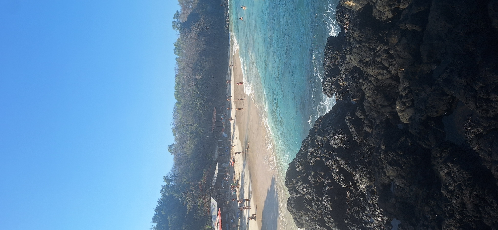 This is a vertical, wide-angle, eye-level shot of a tropical beach scene in Manggis, Indonesia. In the foreground, dark, textured volcanic rocks fill the bottom third of the frame. Beyond the rocks, a narrow strip of sandy beach stretches across the middle ground, with the bright blue ocean lapping at its edge. Several people, dressed in swimwear, are visible walking, sitting, and standing on the beach. Some are near a row of small, open-air structures with thatched roofs, likely beachside stalls or cafes. The ocean itself is a vibrant turquoise and blue, with gentle waves breaking on the shore. The background consists of a dense, green, treed hillside that slopes upwards. The sky is a clear, bright blue with no clouds. The overall impression is a sunny, active beach day in a tropical setting.