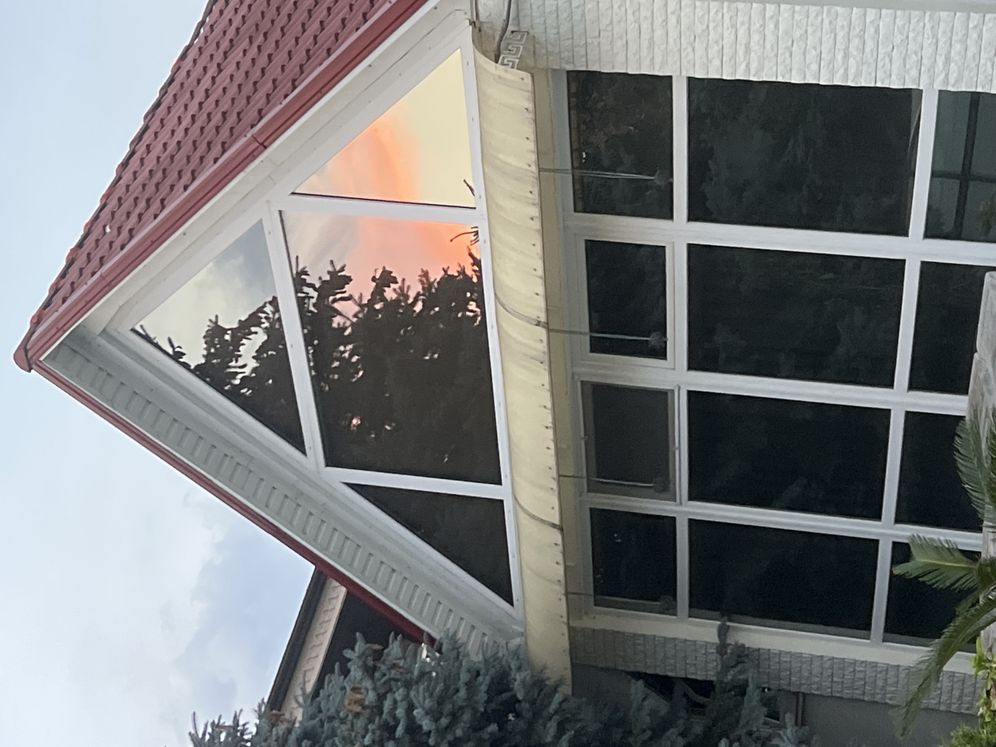The image shows the upper part of a house with a distinctive triangular window, located in the city of Fontanka, Ukraine. The roof is covered with red tiles. The triangular window reflects a sunset with orange and yellow hues, and the silhouette of a tree. Below the triangular window, there is a translucent awning and a larger rectangular window divided into several panes, which are dark, reflecting the interior of the house. A portion of a large blue-green conifer tree is visible on the left side of the image, and a white brick wall with horizontal grooves forms the right side of the house. The sky is overcast with gray clouds.