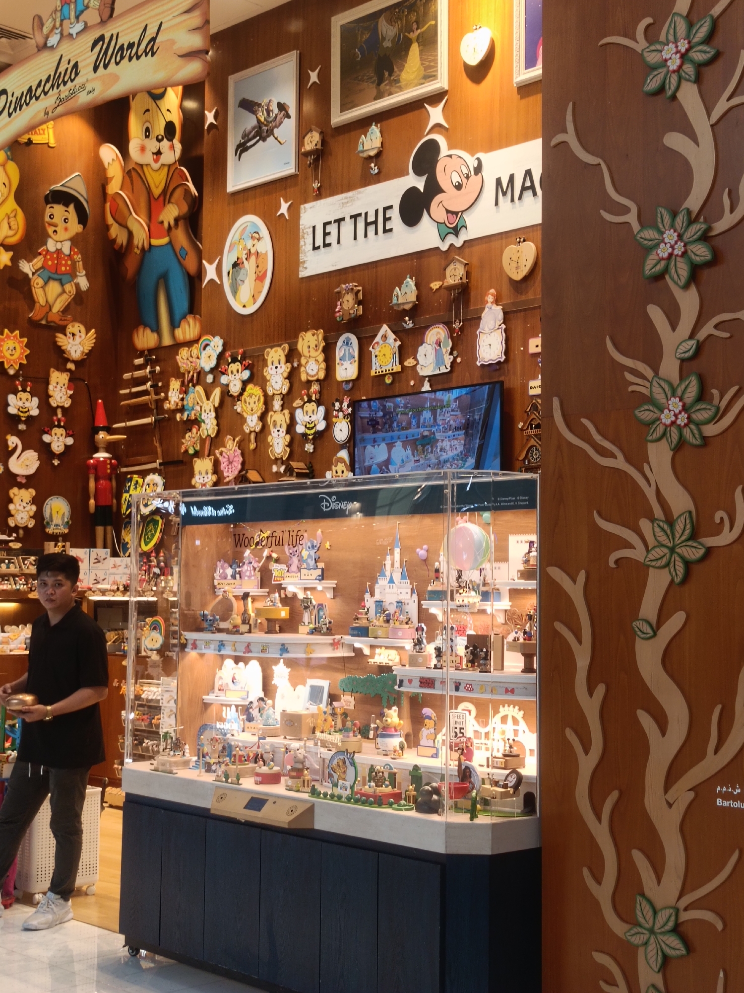 The image shows the interior of a shop, likely a souvenir or toy store, featuring a variety of wooden decorations and merchandise. The shop's walls are adorned with colorful, hand-painted wooden figures, including characters from Pinocchio and Mickey Mouse. There is a large display cabinet filled with smaller wooden figurines and toys, many of which appear to be themed around Disney characters and fairy tales. The overall aesthetic suggests a whimsical and child-friendly environment. The presence of a speed limit sign with "55" indicates an indoor display area, not an outdoor setting. The wooden tree decoration on the right side of the image is a recurring decorative element within the shop. The lighting is warm and inviting, illuminating the details of the merchandise. The man in the foreground, wearing a black t-shirt and dark pants, is facing away from the camera and appears to be interacting with some items.