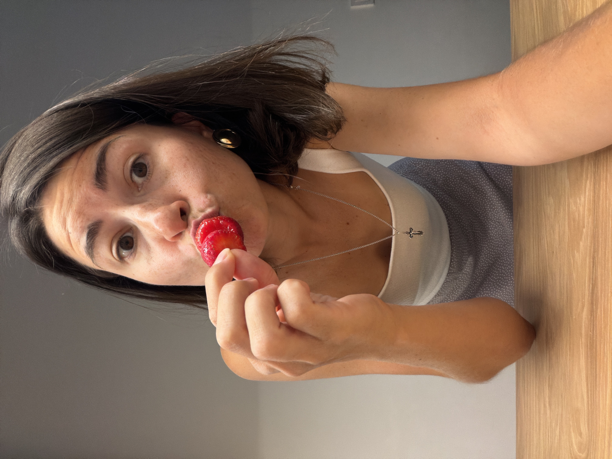 A woman is positioned at the top of the frame, her head tilted downwards. She has short, dark hair, fair skin, and is wearing a silver necklace with a cross pendant. She is in the process of eating a strawberry, which she holds in her left hand close to her mouth. She is also wearing a white tank top and a blue patterned skirt. A wooden table is visible at the bottom of the frame on the right side and a gray wall is to the left. The setting of the image is not in Budva, Montenegro. There is no information that suggests the location.