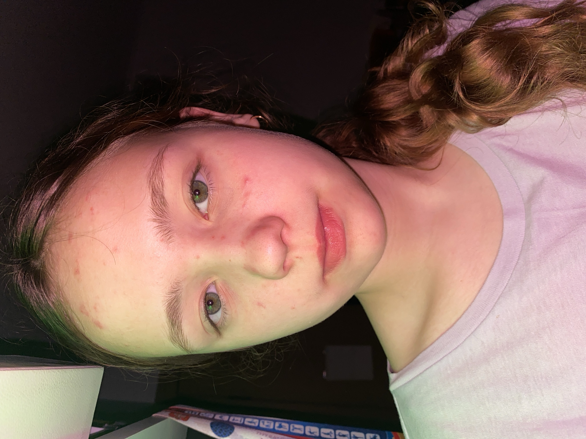A young person with fair skin, light brown curly hair, and green eyes is shown in a close-up portrait. The person is wearing a light-colored t-shirt. The background is dark. The lighting appears to be uneven, with a slight green cast on the left side of the face. The location is Unknown, Unknown.