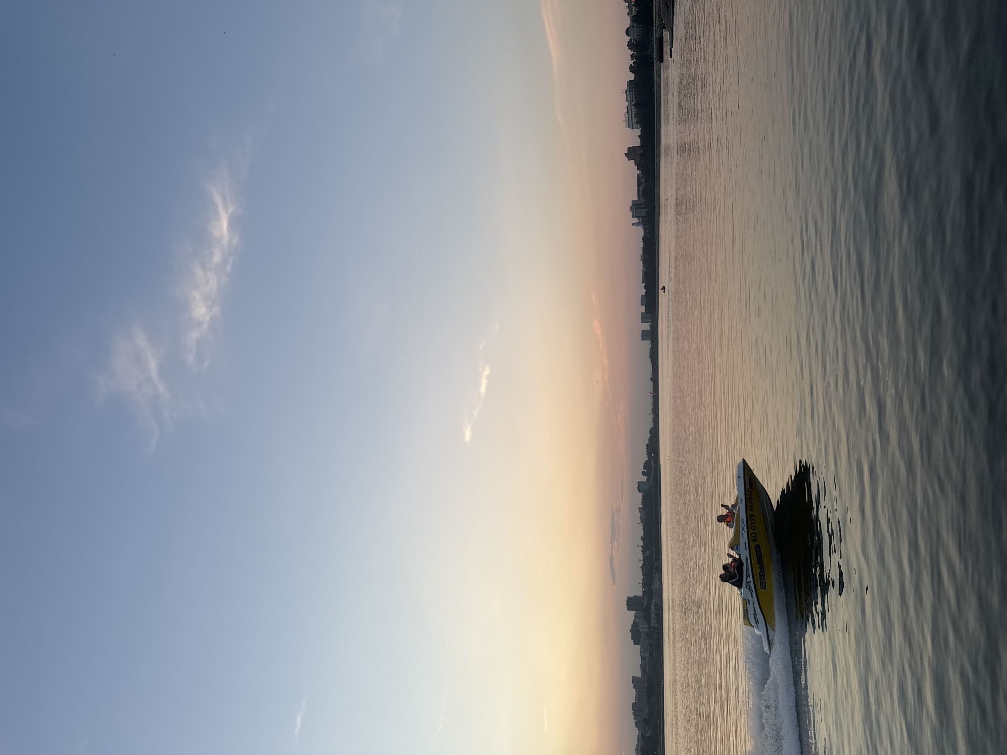 At sunset, a yellow and white speedboat carrying at least three individuals cruises across a large body of water, leaving a white wake. The boat is emblazoned with the name "SEA DOO." In the background, a city skyline with numerous buildings is visible along the distant shore. The sky above transitions from a pale blue at the top to soft hues of orange and pink near the horizon, indicating the time of day. The water is calm, reflecting the colors of the sky. This scene is captured on the Angara River in Irkutsk, Russia.