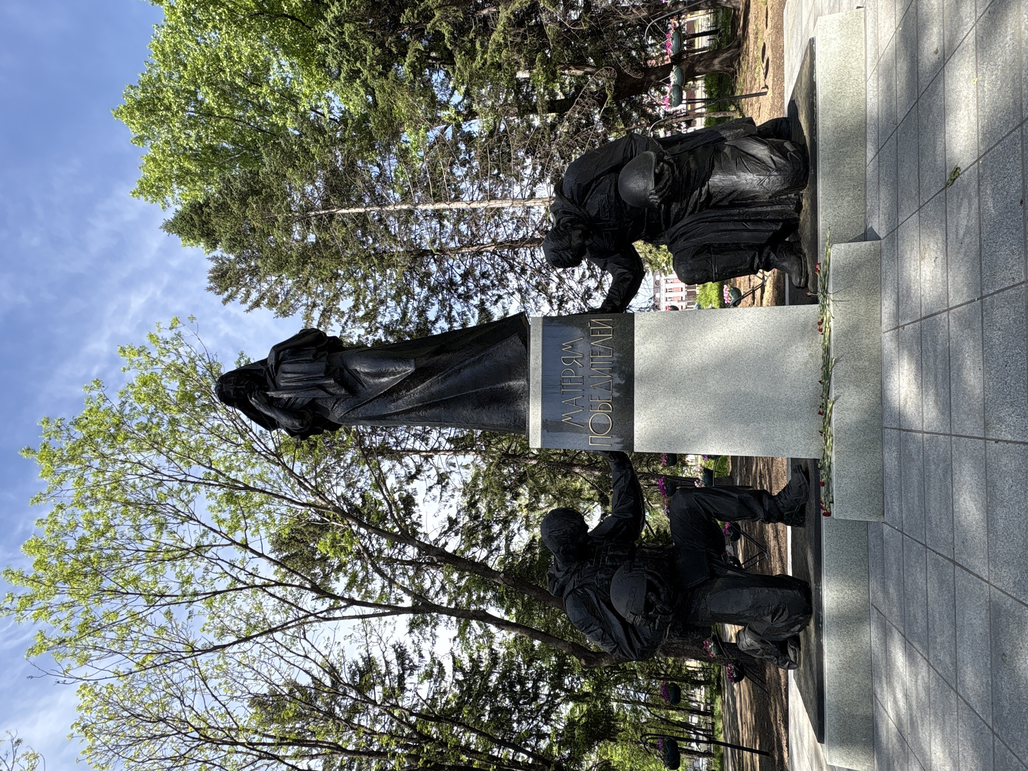 The image shows the Motherland Monument in Birobidzhan, Russia. It depicts a somber, draped female figure standing on a gray plinth next to two kneeling soldiers in dark uniforms and helmets. The plinth bears inscription in Cyrillic, "МАТЕРЯМ ПОБЕДИТЕЛЕЙ," translating to "To the Mothers of the Victors." The statues are dark in color, possibly bronze or another dark metal. The surrounding environment includes trees with green foliage and a partly cloudy sky. The monument stands on a paved area, likely a plaza or park.
