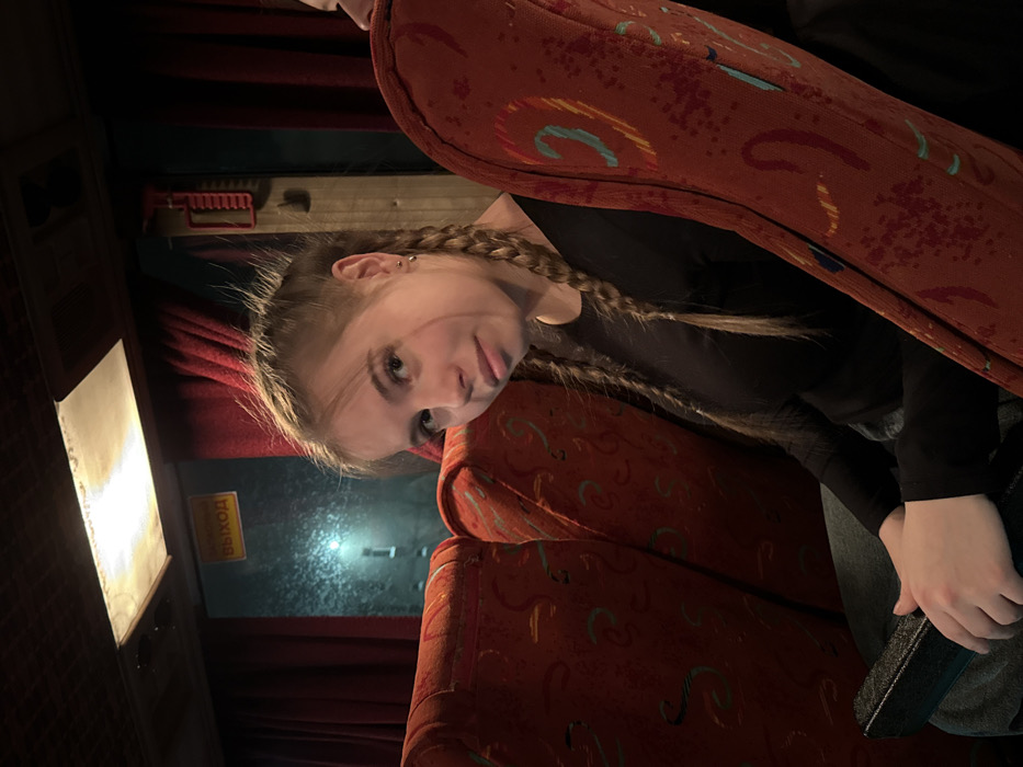 The image captures the interior of a passenger bus, located in Bolshaya Sosnova, Russia. A young light-skinned female with light brown hair styled in two braids is seated in the foreground, facing slightly towards the camera. She wears a black long-sleeved shirt and blue jeans, with small stud earrings visible in both earlobes. Her hands are clasped over a dark, textured, rectangular object resting in her lap. The upper profile of another passenger's head is partially visible on the far right of the frame, behind the primary subject. The bus interior features multiple rows of red upholstered seats, adorned with a faded, abstract patterned fabric. A window is visible behind the primary subject, framed by red curtains, and appears frosted or wet, obscuring the view of the external environment except for a distant, bright streetlight. A yellow sign with the Cyrillic word "ВЫХОД" (EXIT) is illuminated above the window. A discolored rectangular overhead light fixture is present on the ceiling, and a red plastic lever or handle is mounted on the wall to the right of the window. The overall lighting suggests low light conditions, possibly nighttime.