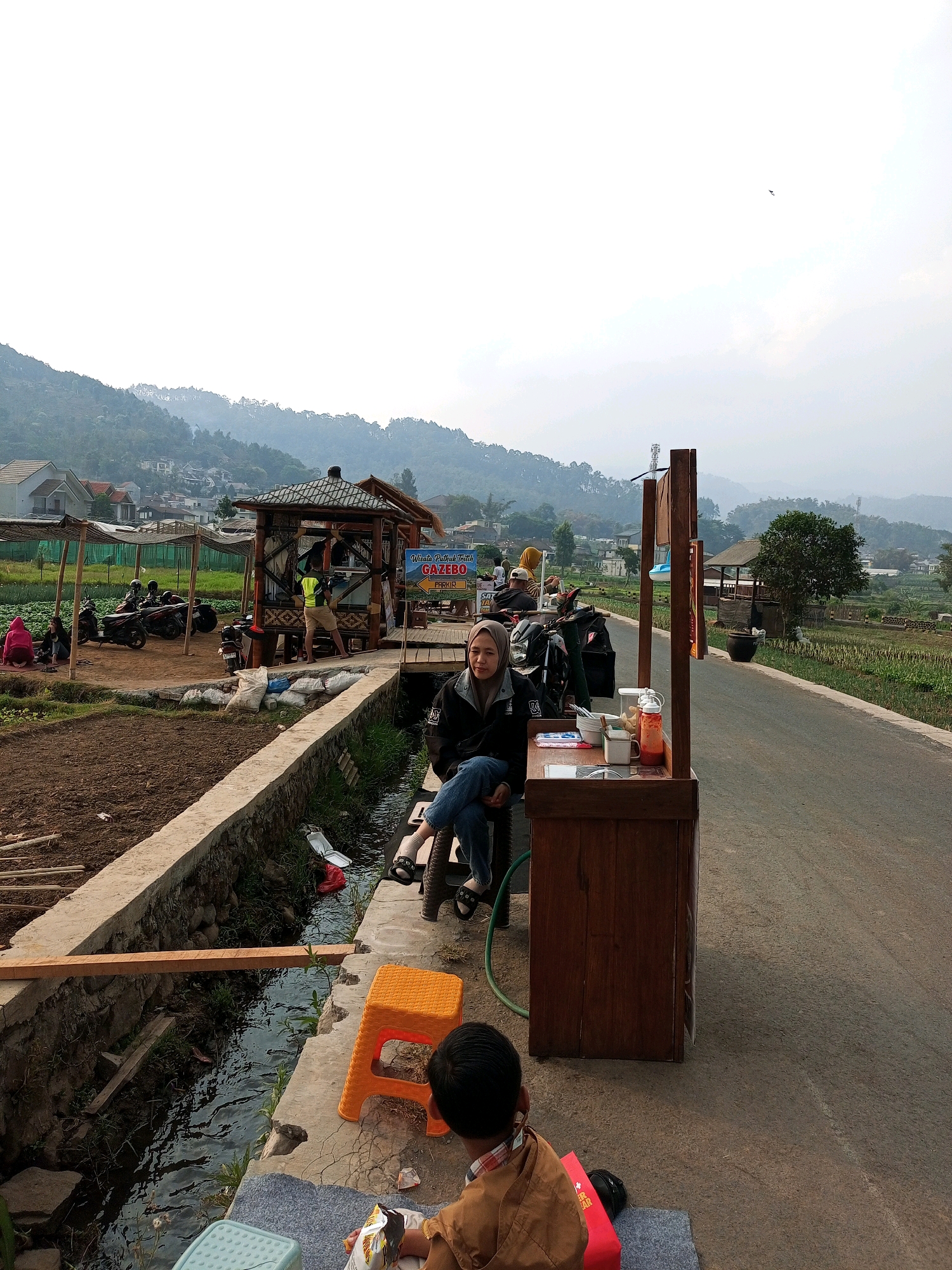 This is a landscape photograph taken in Batu, Indonesia, likely a rural or semi-rural area with rolling hills in the background. The image depicts a roadside scene with several elements. In the foreground, to the right, is a rustic wooden cart or stall displaying various food or drink items and condiments, including bottles of sauce and cups. A woman wearing a hijab and dark jacket is seated on a dark chair next to the cart, looking towards the camera. To the left of the cart, a small orange plastic stool and a small child wearing a brown jacket are visible. A narrow concrete drainage ditch runs alongside the road, with water flowing in it. There's a wooden plank laid across the ditch.
Further back, a small, traditional gazebo structure with a thatched roof is visible, which appears to be part of a tourist attraction, as indicated by a sign that reads "Wisata Petik Tirth... GAZEBO PARKIR" (Tourism Pick Fresh... GAZEBO PARKING). Several motorcycles are parked near the gazebo, suggesting a place for visitors to stop. In the middle ground, there are cultivated fields or gardens with dark soil. In the background, the landscape rises into mist-covered hills dotted with houses and trees. The sky is overcast, contributing to the soft, diffused light. The overall atmosphere suggests a relaxed, possibly tourist-oriented stop in a scenic natural setting.