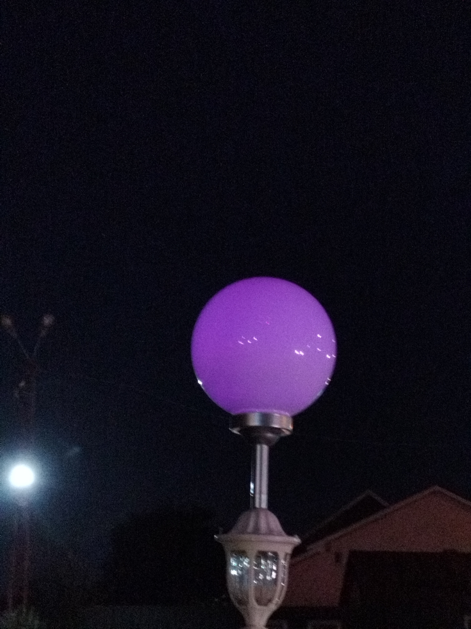 The image captures a close-up view of a purple-glowing spherical light fixture mounted on a lamppost at night. The background is dark, suggesting it is nighttime, with a hint of a building's roofline visible in the lower right quadrant. The light fixture itself is luminous and emits a soft, diffused purple light. The lamppost has a metallic stem and a decorative base. In the upper left portion of the image, a faint silhouette of what appears to be another lamppost or street light can be observed. The overall scene appears to be an outdoor setting in a city. No people are visible in the image. Based on the information provided, this image is located in Bucharest, Romania.