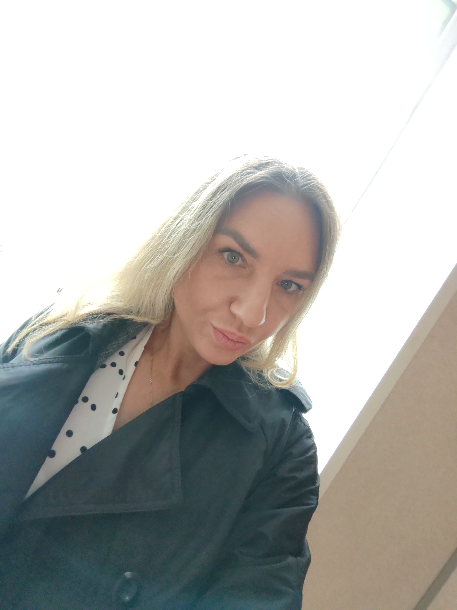 The image is a medium close-up selfie of a woman, taken from a low, upward angle. The primary subject is a woman with long, straight, blonde hair that cascades around her shoulders. She has light-colored eyes, appearing green, and a fair complexion. She is looking directly at the camera with a somewhat confident or playful expression, characterized by slightly pursed lips.
She is dressed in a dark-colored, possibly black or navy, jacket or coat with a collared neckline. Underneath, a white blouse or top with black polka dots is visible. A thin, delicate, gold-colored chain necklace is also seen around her neck.
The background is largely overexposed and bright white, suggesting a powerful light source, most likely a large window, behind her. This strong backlighting creates a bright halo effect around her hair. On the right side of the frame, a segment of a light brown or beige wall or interior surface is visible, indicating an indoor setting. The intense natural light implies the photograph was taken during daytime. There is no discernible text within the image.