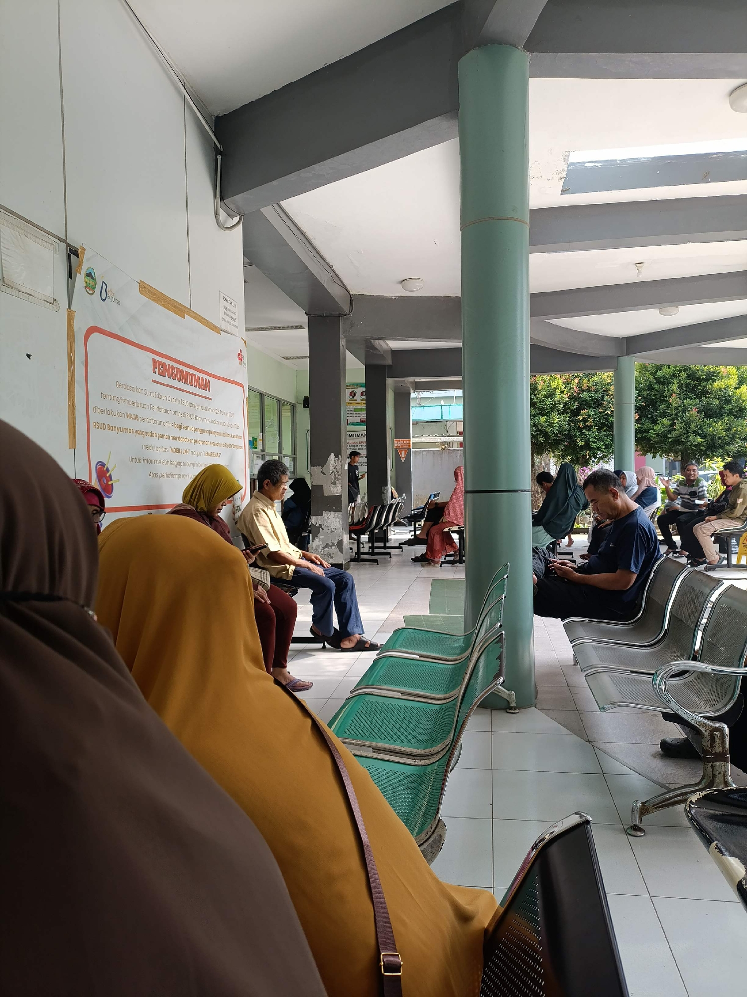 The image depicts a waiting area, possibly at a public health facility, in the city of Kejawar, Indonesia. A diverse group of individuals are seated on rows of metal benches. The architecture features a covered walkway with supporting pillars and a tiled floor. The lighting suggests it is daytime, with natural sunlight illuminating the scene, casting shadows. In the background, greenery and other buildings are visible. A poster with Indonesian text is displayed on a wall. The people are dressed in casual clothing, some wearing headscarves. The overall atmosphere appears to be one of quiet waiting.