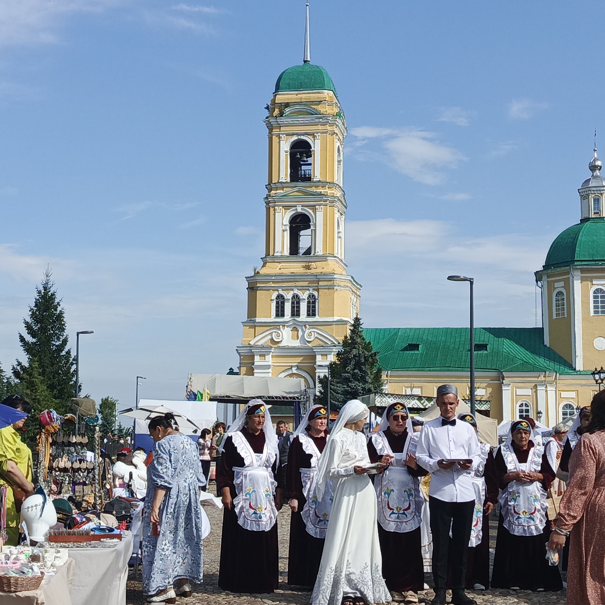 In Nikolo-Beryozovka, Russia, a group of people dressed in traditional attire is gathered outdoors. A prominent yellow bell tower with a green dome stands in the background, adjacent to a building with a green roof. Several women are wearing dark dresses with white aprons adorned with colorful embroidery, and headscarves. One woman in the center is wearing a white wedding dress and veil. A man in a white shirt and black bow tie is standing with them. The group appears to be participating in a cultural event or festival, with stalls selling various items visible to the left. The sky is blue with scattered clouds.