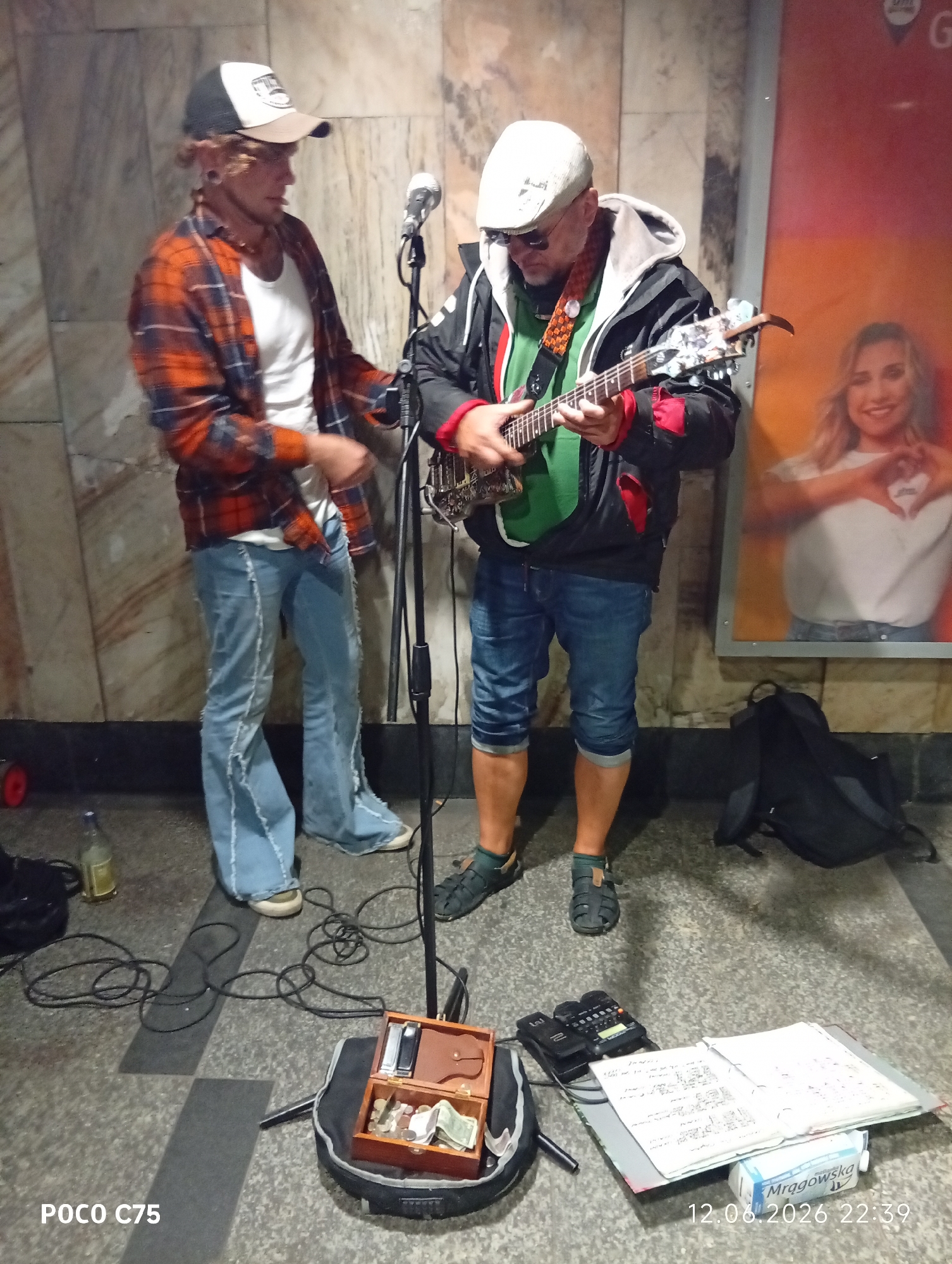 The image depicts two men performing music in what appears to be a subway station or underground walkway in Wrocław, Poland, as indicated by the timestamp and the brand name "Mrągowska" visible on a small carton.
In the foreground, on the left, a younger man with ear gauges and wearing a plaid shirt over a white t-shirt and distressed denim jeans is standing and looking down, possibly at the microphone stand or other equipment. He has a baseball cap on.
To the right, an older man wearing a white cap, sunglasses, a green t-shirt, and a dark jacket with red accents is playing a small electric guitar. He is also wearing denim shorts.
In front of them on the ground, there is a collection of musical equipment. A microphone stand is centrally placed. There is a wooden box open and filled with coins and some banknotes, suggesting it is a donation box for their performance. A black bag is visible behind the older musician. Further to the right, a black electronic device, possibly a loop station or amplifier, is connected to cables, and an open notebook or sheet music is spread out. A bottle with liquid is on the floor near the younger man.
The background shows a tiled wall, typical of public transportation infrastructure. A large poster featuring a smiling woman is visible on the right side of the frame. The floor is made of textured pavement. The overall impression is a street performance taking place in an urban, possibly underground, public space.