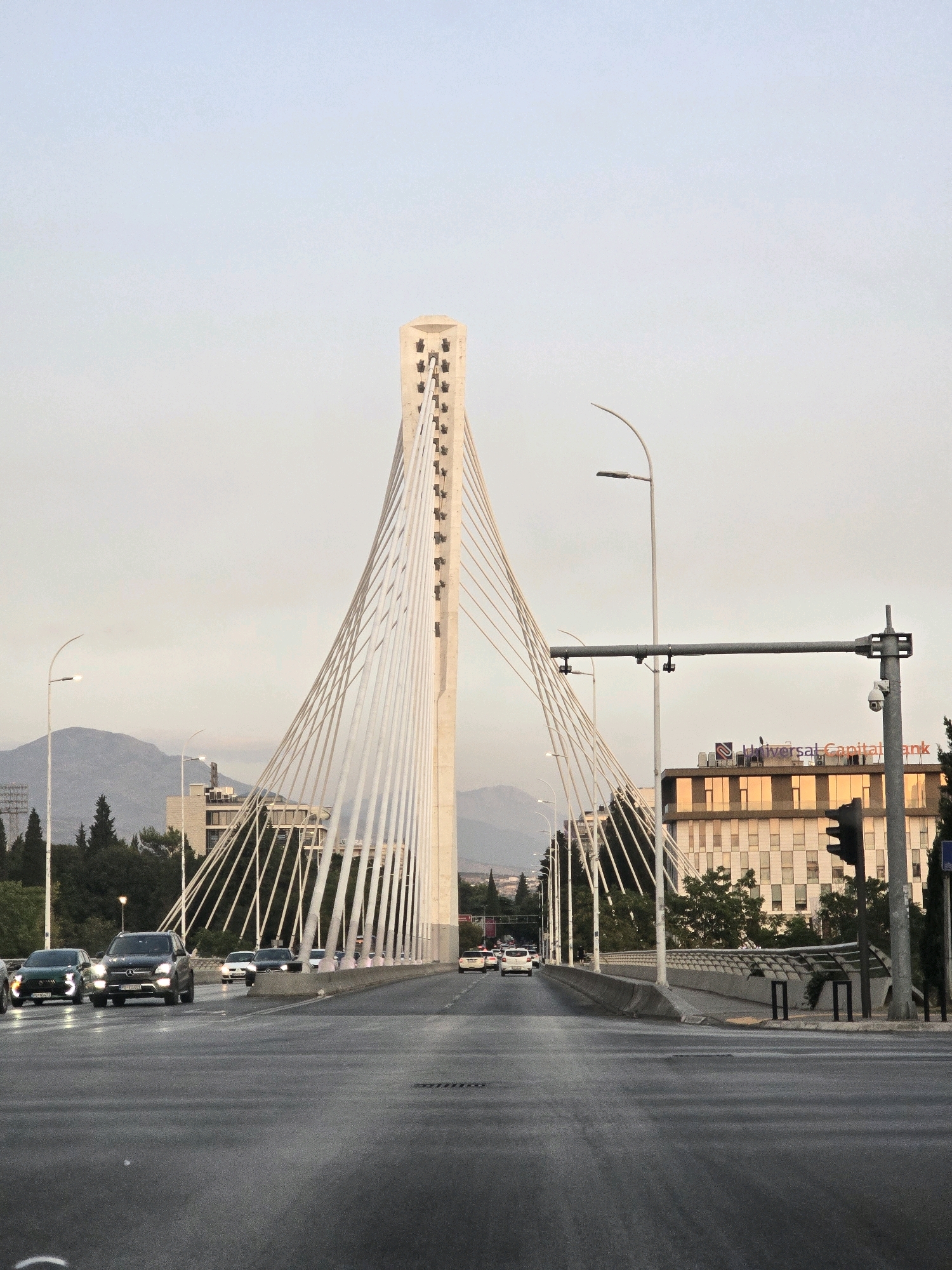 This is a medium shot, eye-level, vertical photograph taken from a moving vehicle looking forward along a paved road towards a suspension bridge. The bridge has a tall, narrow, light-colored pylon from which numerous white cables descend to support the road deck. On the right side of the road, a modern building with reflective windows bears the signage "Universal Capital Bank." A traffic light is visible in front of the building. Several cars are traveling on the bridge in both directions. Trees and distant mountains are visible in the background. The sky is clear and pale, suggesting it is either early morning or late afternoon. This is a view of the Millennium Bridge in Podgorica, Montenegro.