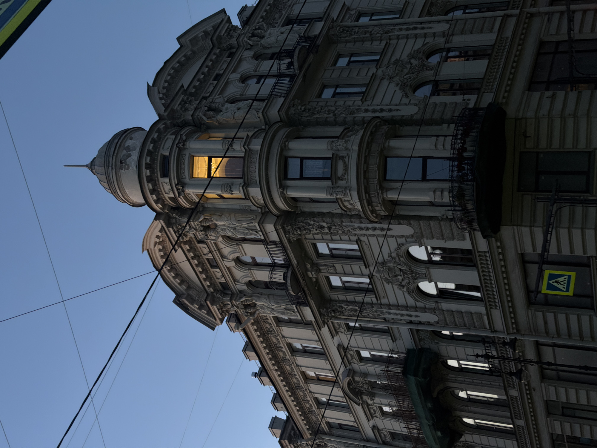 The image captures a grand, ornate building in Saint Petersburg, Russia, at dusk. The architecture is characterized by intricate detailing, including sculptures adorning the facade, balconies with wrought-iron railings, and decorative moldings around the windows. A prominent turret, topped with a small dome and spire, juts out from the corner of the building. One window in the turret is illuminated from within, casting a warm, golden glow against the darkening twilight sky. Utility wires crisscross the sky above the building, a common sight in urban environments. In the foreground, a pedestrian crossing sign with a yellow triangular warning symbol is visible, indicating the presence of a street. The overall impression is one of historical grandeur and urban atmosphere.