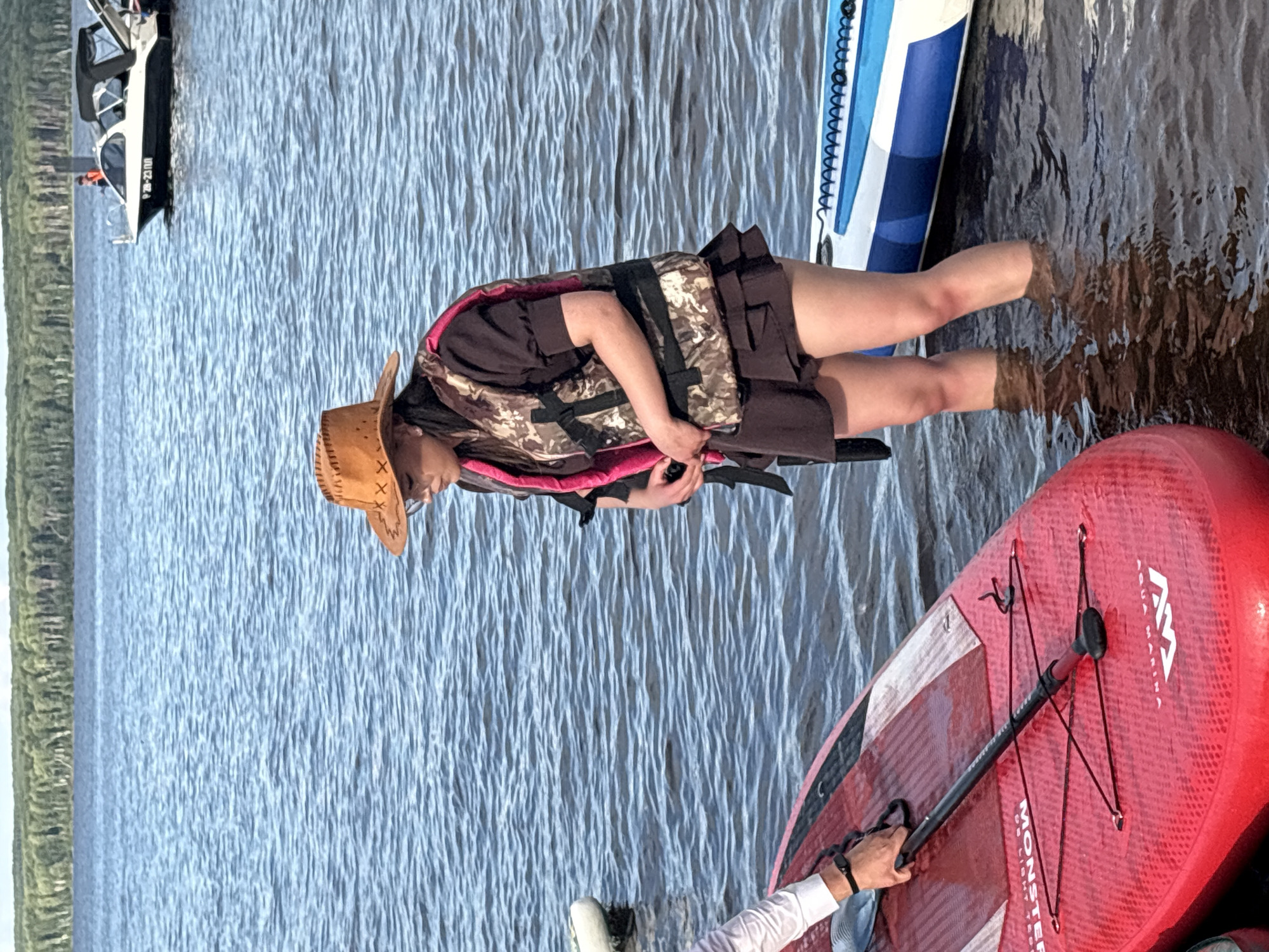 A young woman wearing a cowboy hat and a camouflage life vest stands in the water at the edge of a lake. She is holding a paddle. In the foreground, there is a red paddleboard and a blue and white paddleboard. In the background, a small boat with a black canopy is visible on the water, with a treeline on the horizon. The setting is likely a recreational area near Chaikovsky, Russia, given the presence of watercraft and natural scenery typical of the region.