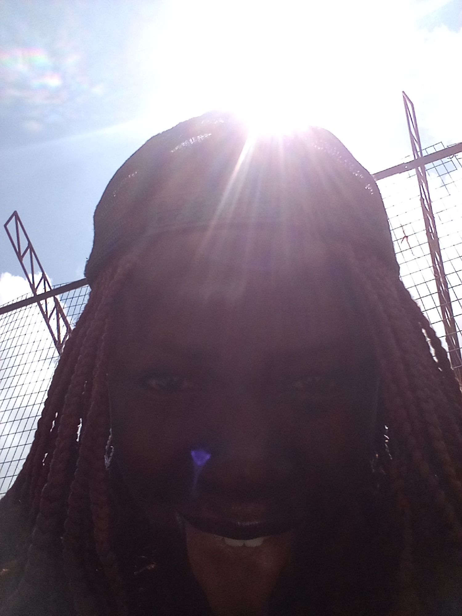 The image depicts a close-up, low-angle selfie of a young Black woman with braided hair, wearing a dark-colored cap and a visible earring. The sun is directly overhead, causing lens flare and obscuring parts of her face. Behind her, a chain-link fence with red metal supports is visible, against a bright sky with scattered clouds. The scene appears to be outdoors in an urban setting, possibly New Stadium, Nigeria, given the context. The woman's mouth is slightly open, showing teeth, and she is looking towards the camera.