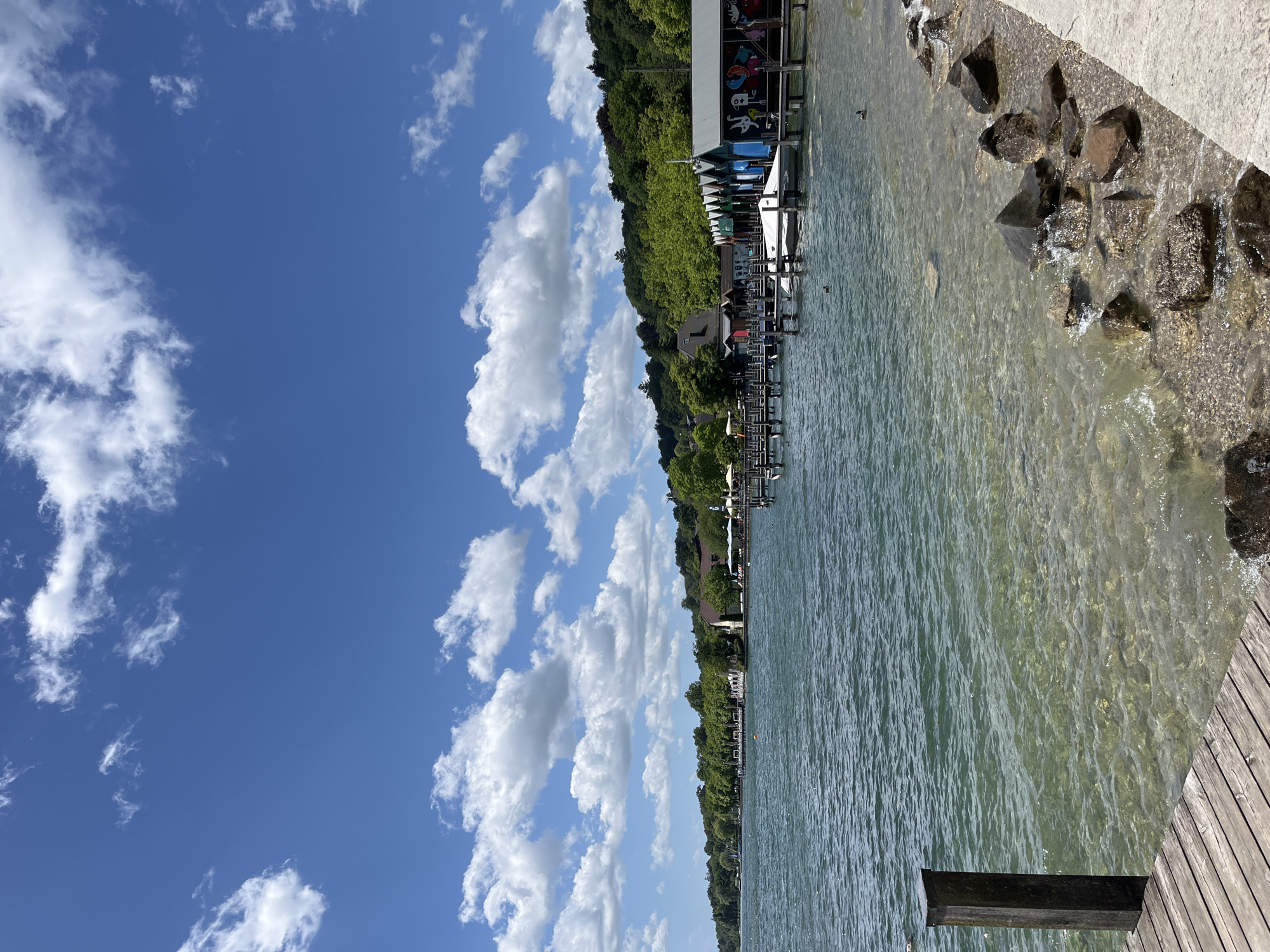 The image captures a scenic view of the Starnberger See in Germany on a bright, partly cloudy day. In the foreground, a wooden pier extends into the clear, shallow water, revealing a pebbly lakebed. To the right, a stone embankment lines the shore, with several dark rocks partially submerged in the water.
The middle ground features a wooden dock with multiple boathouses, each painted in vibrant colors and adorned with eclectic artwork. Some boathouses are closed, while others are open, revealing boats stored inside. A few ducks are visible in the water near the dock.
The background showcases a lush, tree-covered hillside that slopes down towards the lake. A row of buildings, possibly restaurants or residential structures, are nestled among the trees along the shoreline. The sky is a brilliant blue, dotted with fluffy white clouds. The overall atmosphere is one of a peaceful summer day by the lake.