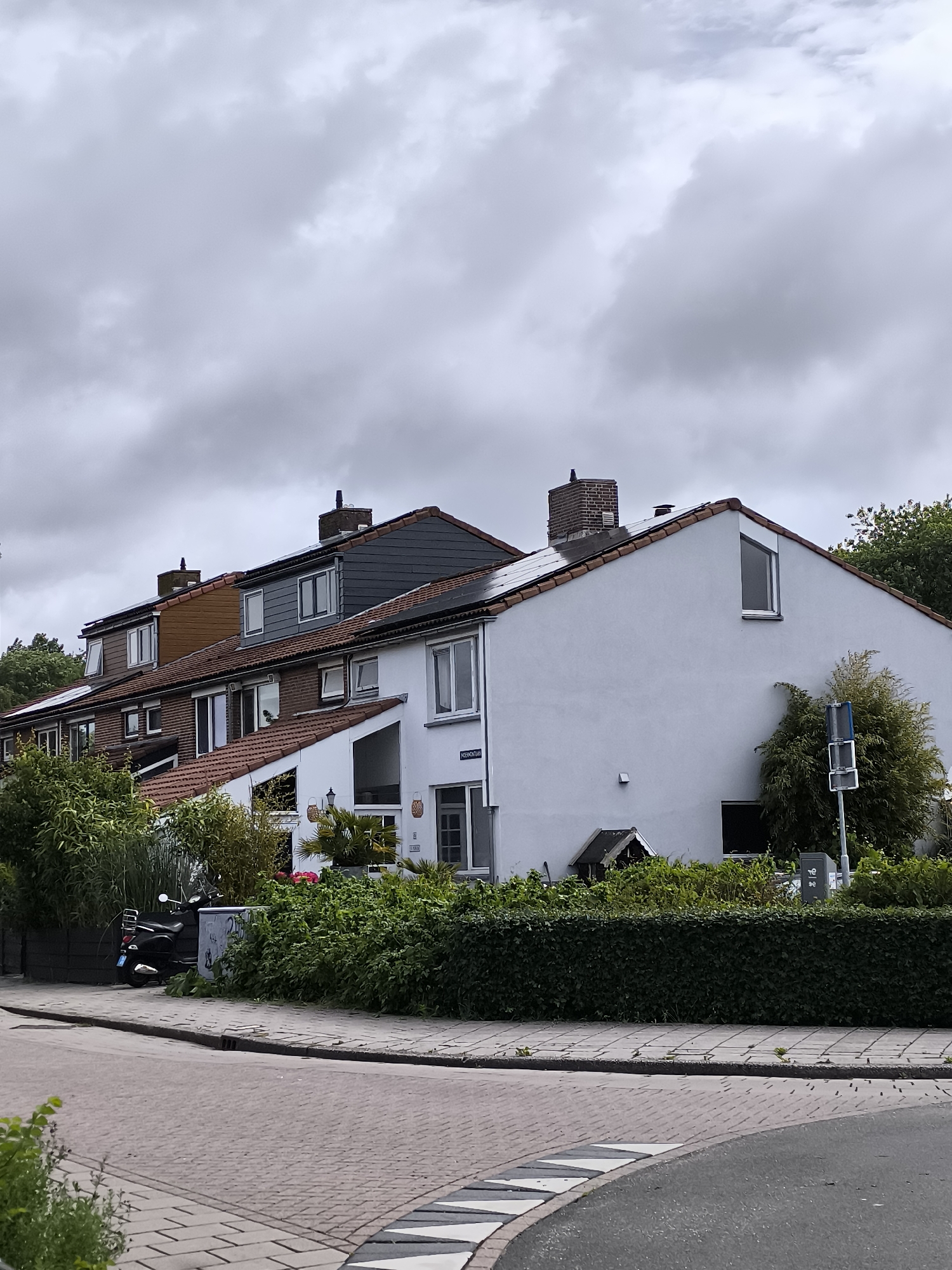 The image depicts a residential street scene in Alkmaar, Netherlands, under an overcast sky. A row of terraced houses with red tile roofs and various wall materials, including white render and brick, are visible. The houses have multiple windows and chimneys. Lush green hedges and trees line the front of the properties. A black scooter is parked on the sidewalk next to a dark fence. The street in the foreground is paved with interlocking bricks and asphalt, with a visible speed bump. A street sign with text is affixed to one of the houses.