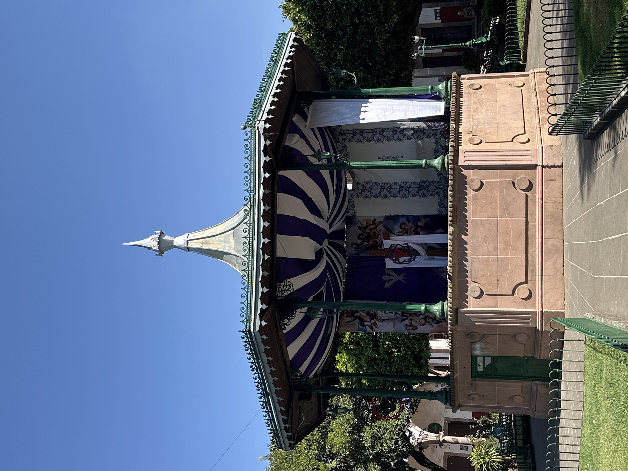 The image depicts a bandstand in the main plaza (Plaza Principal) of Quiroga, Michoacán, Mexico. The structure has a stone base with green metal columns supporting a roof decorated with white and purple striped fabric. There are decorative murals or paintings hanging inside the bandstand. The sky is clear and blue. Green foliage and a green metal fence surround the bandstand. A paved walkway and grass are visible.
