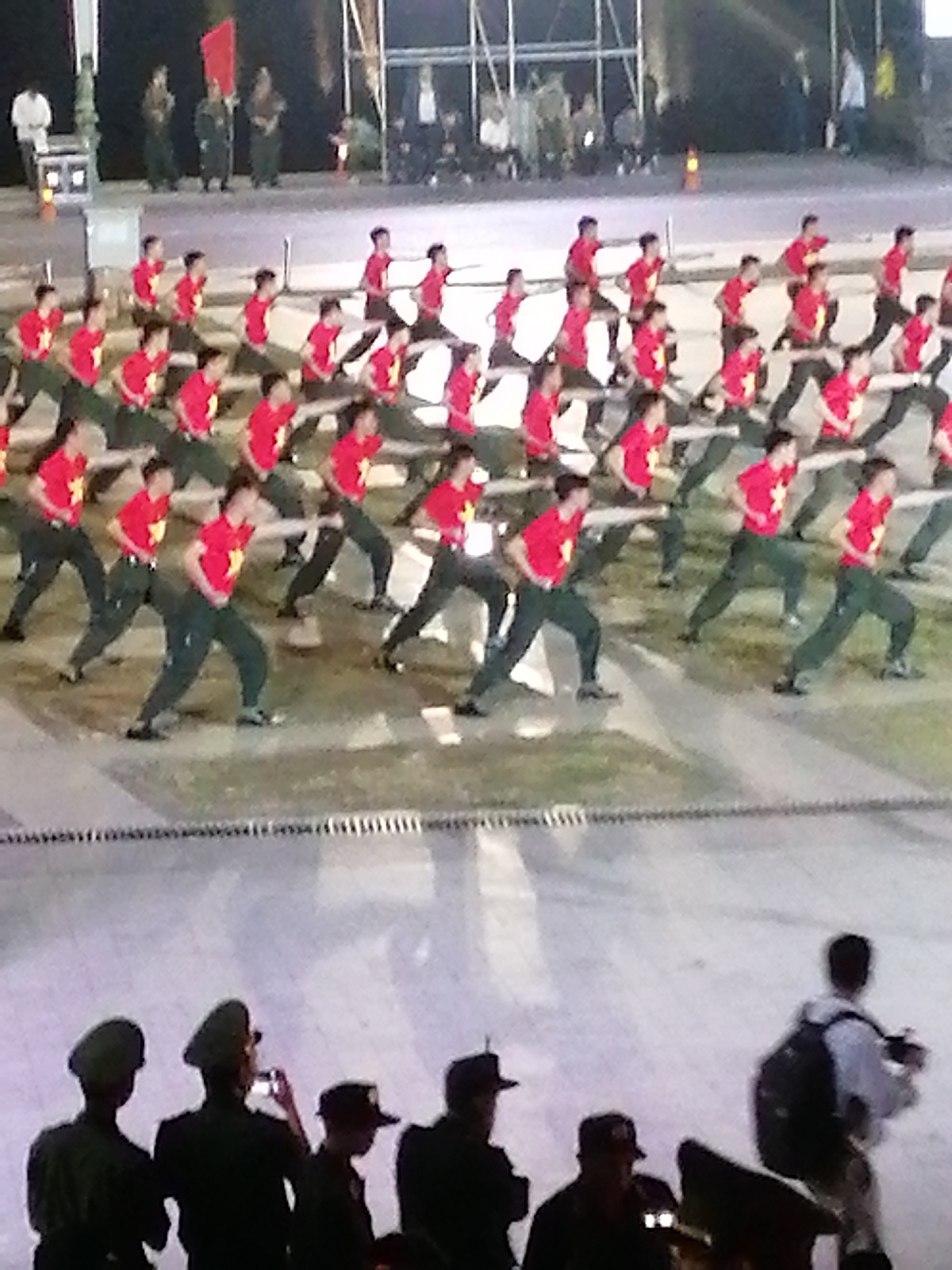 In Da Lat, Vietnam, a group of individuals in matching red shirts and dark pants perform a synchronized dance or exercise routine on a paved outdoor surface. The participants are arranged in rows and columns, with their bodies angled in various positions. In the background, other individuals, some wearing uniforms and hats, observe the performance. The scene appears to be an organized event, possibly a cultural performance or a public demonstration. The lighting suggests it is either dusk or nighttime.