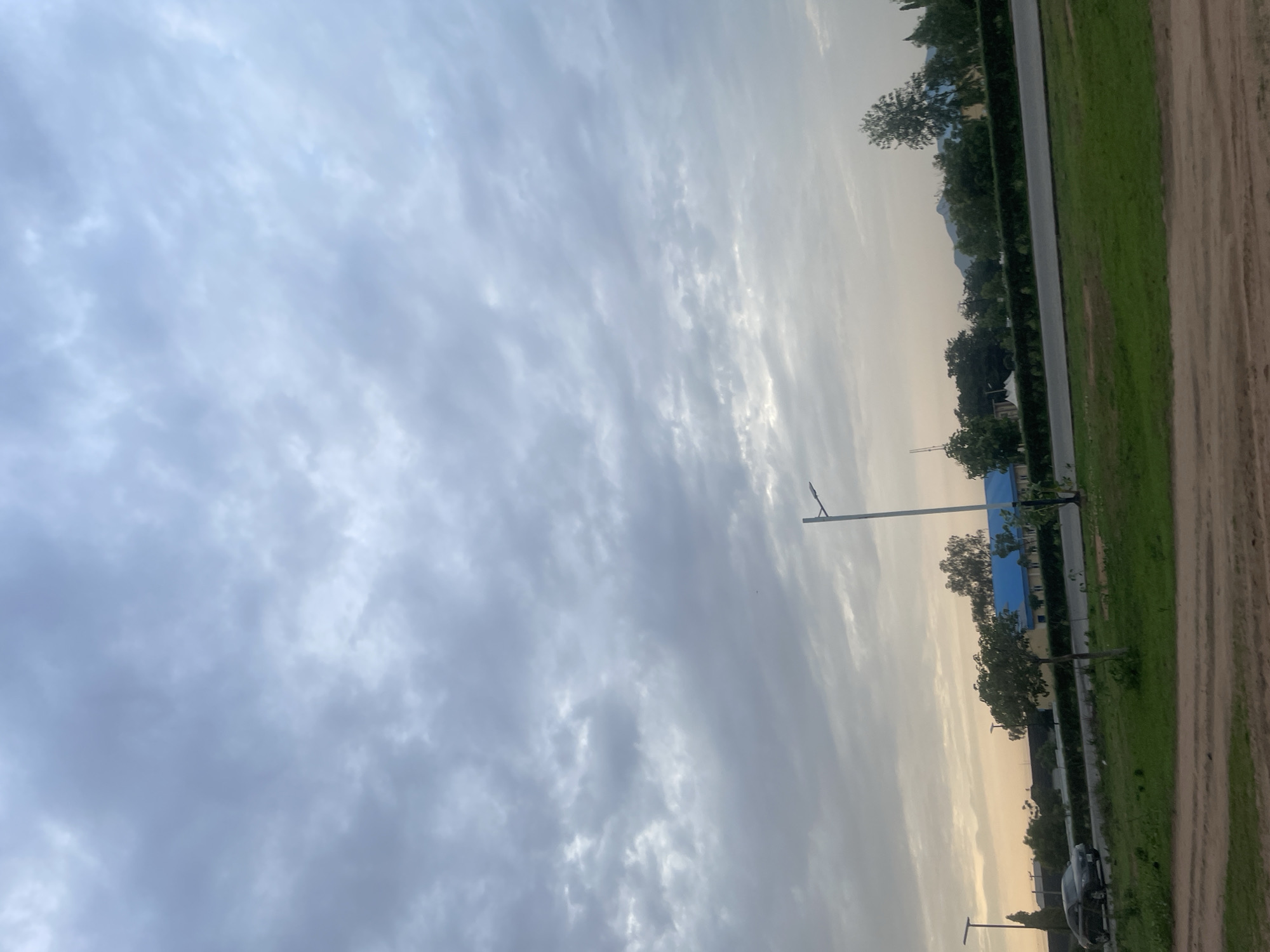 The image depicts an outdoor scene, likely in Jalingo, Nigeria, with a focus on the sky and its surroundings. The sky is filled with a mixture of clouds, with some areas appearing brighter, suggesting either sunrise or sunset. The lighting is soft and diffused, indicating a possible overcast or twilight condition.
The lower portion of the image reveals a landscape that includes a road, a grassy area, and some trees. A light pole with a fixture is visible, suggesting an urban or semi-urban environment. Further in the distance, buildings and more trees can be seen, along with what appears to be a mountain range in the far background. A dark-colored vehicle is parked by the side of the road. The ground in the foreground is a textured, sandy or dirt path.
No people are visible in the frame. The activity, if any, is not discernible. The overall mood is calm and serene, possibly reflecting the quietness of either early morning or late evening. The time of day is suggested to be around dawn or dusk due to the sky's luminosity and the soft light. The weather appears to be cloudy but not necessarily raining. The location cues, such as the vegetation and the distant mountains, are consistent with a Nigerian setting. There is no visible text in the image.