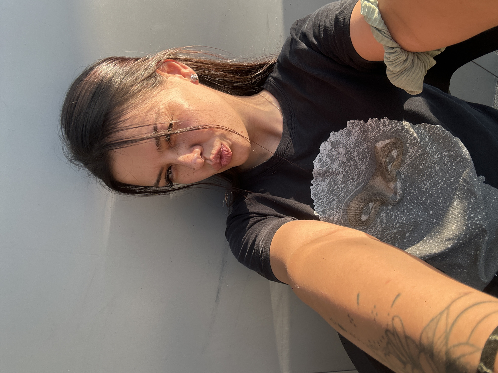 A young woman with dark hair is captured in a selfie outdoors, leaning against a gray wall. She is wearing a black t-shirt with a graphic print of a face. The sunlight casts a warm glow on her skin and hair. Her lips are pursed in a playful expression, and one eye is partially closed, suggesting a wink. She has freckles on her nose and cheeks and is wearing a small, sparkly earring. A scrunchie is visible on her arm. The background is a plain gray wall, likely part of an urban structure in Sliven, Bulgaria.