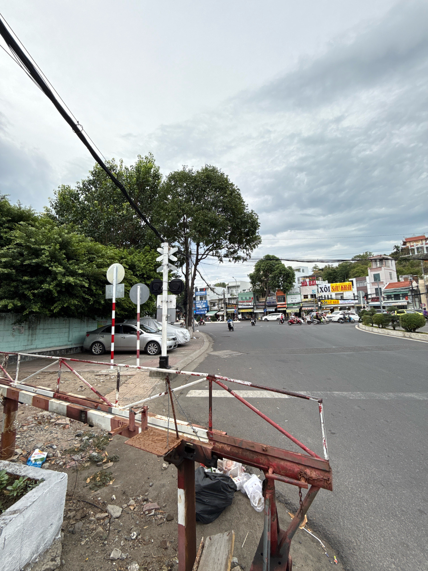 The image depicts a street intersection in Nha Trang, Vietnam, under an overcast sky. In the foreground, a rusty red and white striped barrier is visible, with debris and a black trash bag scattered on the ground beneath it. A gray sedan and a car covered with a tarp are parked along a low, light blue wall on the left side of the street. Further down the street, a traffic signal with red and black lights stands next to a white cross-shaped railway crossing sign. The road is paved, and several motorcycles and a few cars are present in the intersection and on the surrounding streets. Buildings line the sides of the road, many featuring signs with Vietnamese text. One prominent sign displays the text "XÔI SAU KHAI" in yellow. Trees are interspersed throughout the scene, with lush green foliage on the left and sparser trees in the background. The overall impression is a typical urban street scene in Vietnam, with a mix of vehicles, buildings, and everyday elements.