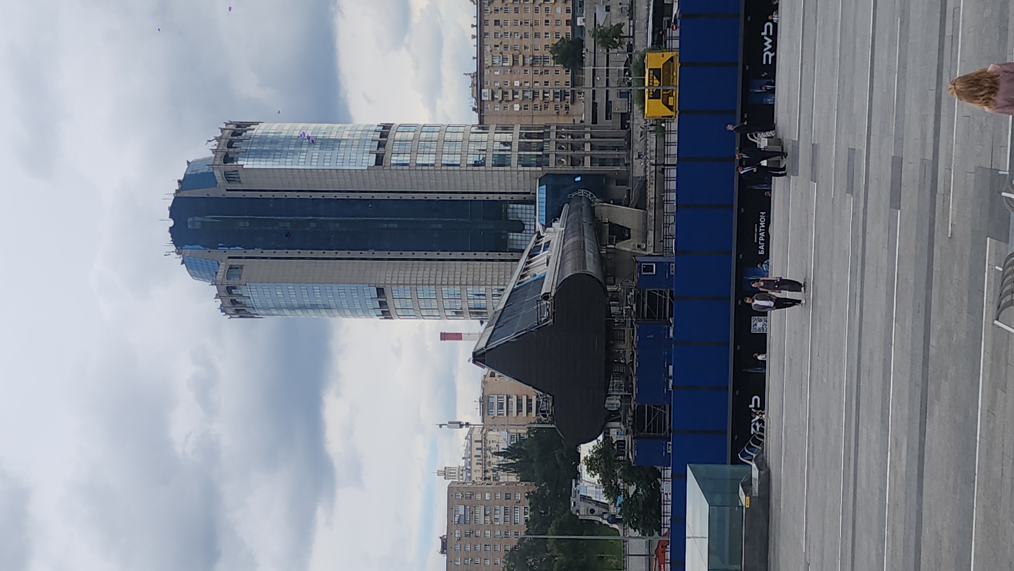 The image shows an outdoor scene in Moscow, Russia. In the background, a tall, modern skyscraper with a rounded top and blue-tinted windows stands prominently. In front of the skyscraper, a large, dark, triangular-shaped object, possibly a section of a train or vehicle, is mounted on a blue platform. Several people are visible in the foreground, walking on a wide, paved plaza. Some are dressed in casual attire, while others are in suits. The sky is overcast with grey clouds. The overall impression is a mix of urban architecture and public space.
