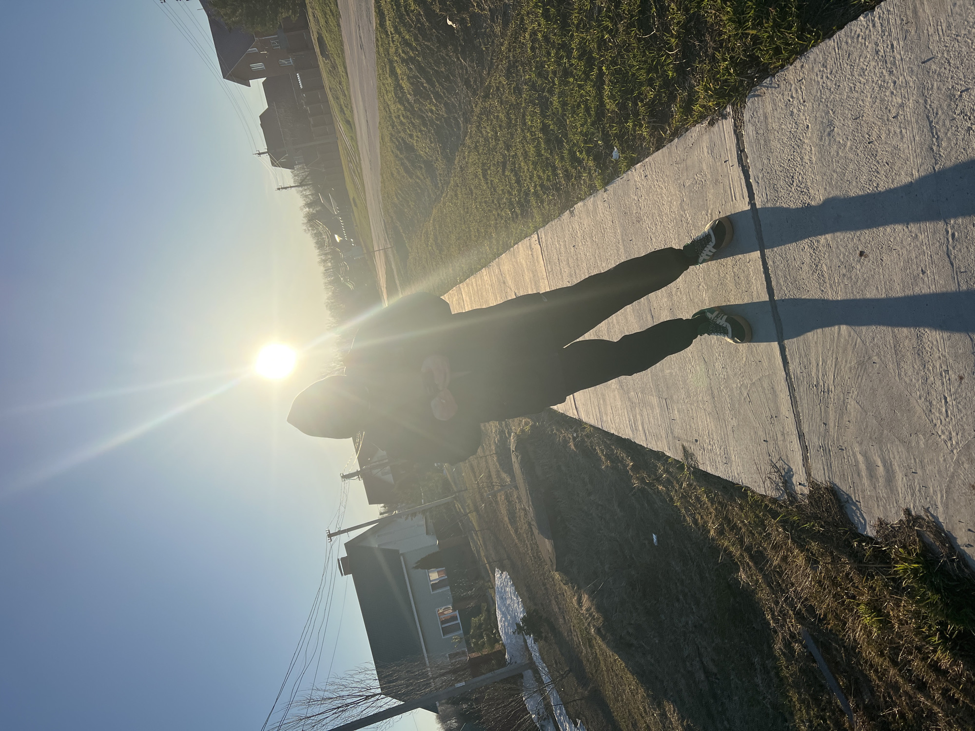 The image depicts a figure standing on a concrete sidewalk next to a grassy area with some dry patches, possibly in Noviy, Russia. The sun is positioned in the background, creating a strong backlight that obscures the figure's features. The figure is wearing dark clothing, including what appears to be a hooded sweatshirt and pants, and dark shoes. The shadow cast by the figure is visible on the sidewalk. Behind the sidewalk and grassy area, houses are visible, indicating a residential area. A patch of snow is visible behind the houses.