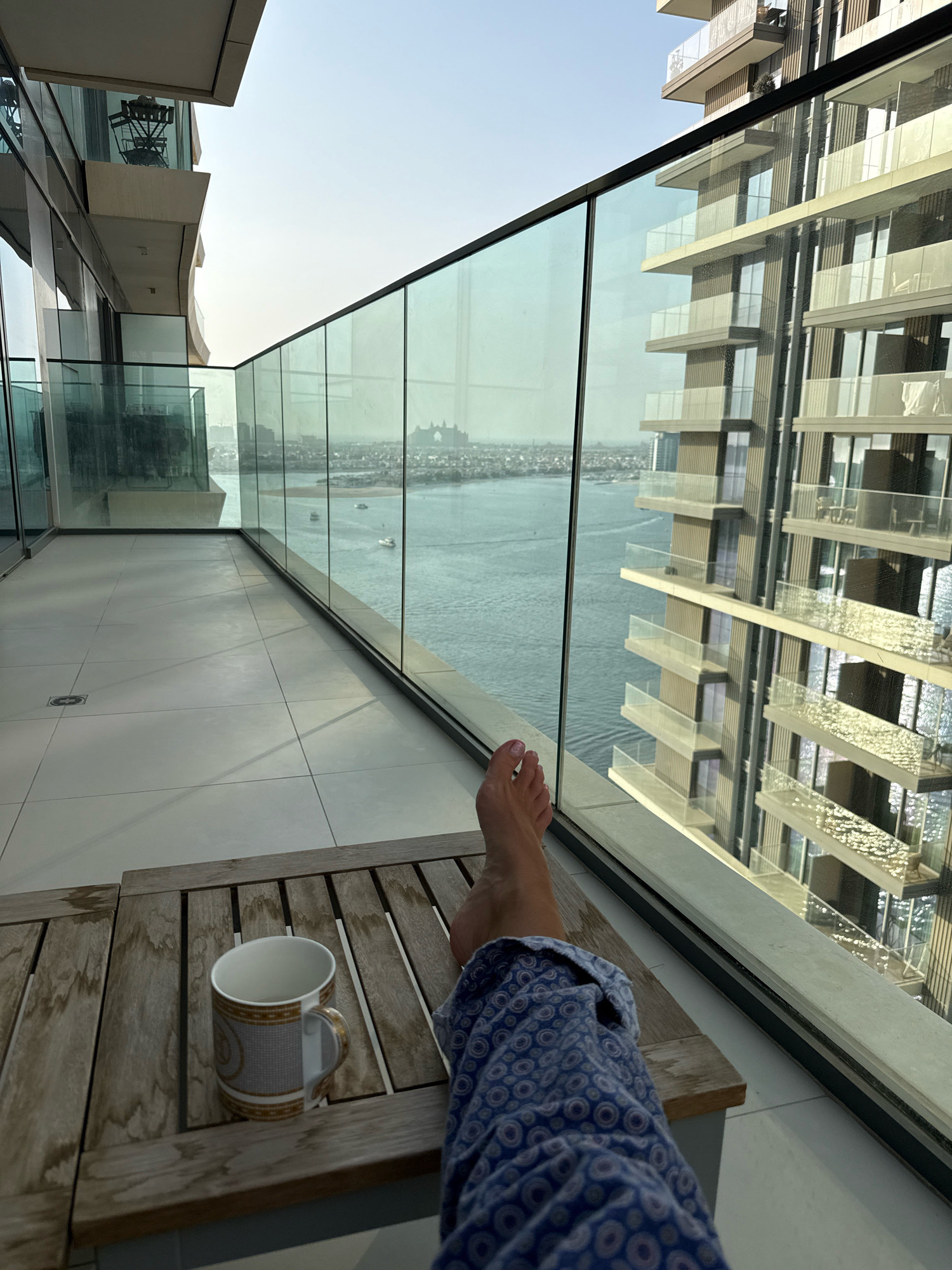 The image depicts a person's feet and lower legs, clad in blue patterned pajama bottoms, resting on a wooden outdoor table. A white mug with a decorative pattern rests on the table. The person is on a balcony overlooking a cityscape in Dubai, United Arab Emirates. To the right, a modern skyscraper with multiple balconies and glass railings is visible. In the distance, a body of water, likely the Arabian Gulf, can be seen with boats sailing on it, and a distant landmass with buildings. The sky is clear and bright, suggesting it is daytime. The balcony floor is tiled in a light grey color.