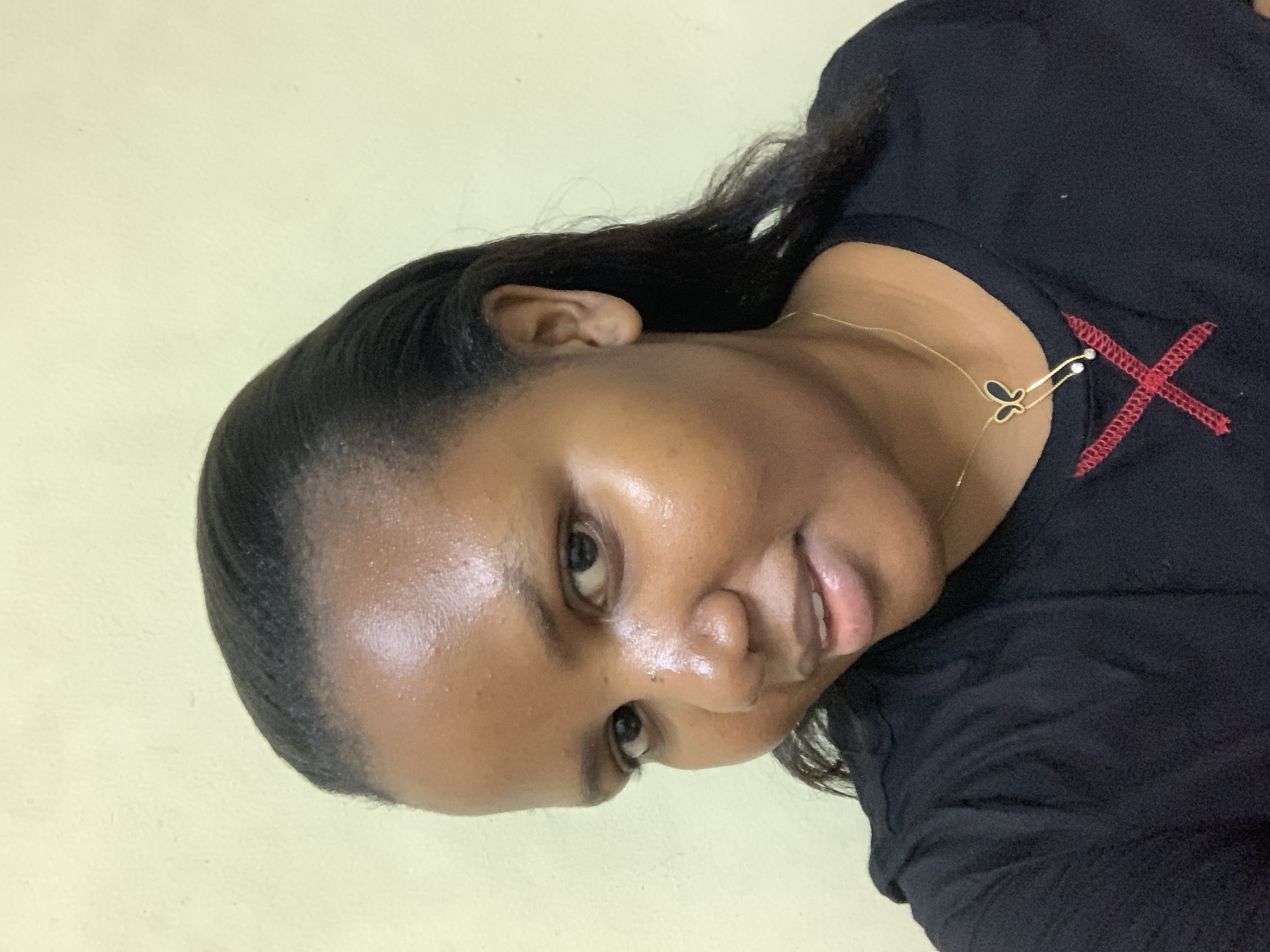 A photograph depicts a woman with dark hair and brown skin. She is wearing a black shirt with a red "X" design stitched onto the shoulder. A delicate gold necklace with a small butterfly charm adorns her neck. The background appears to be a pale yellow wall. The image provides no information to determine if this photo was taken in Dar es-Salaam, Tanzania.