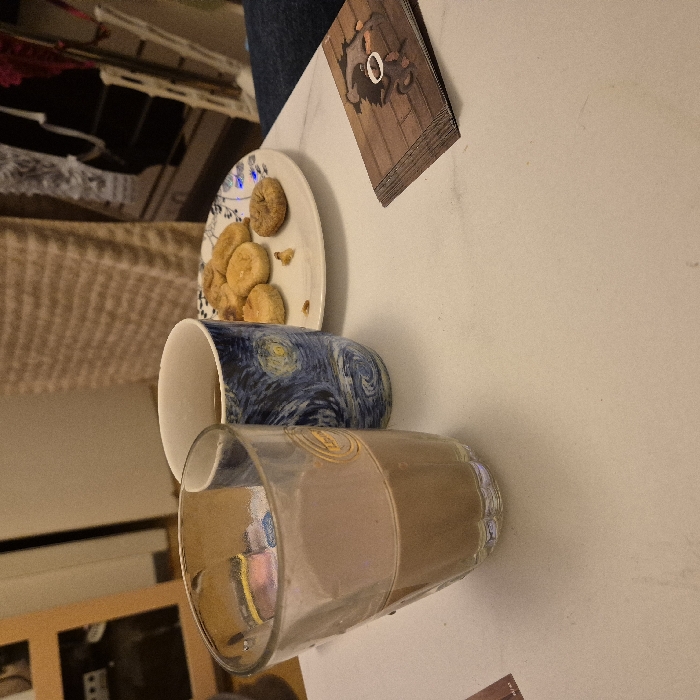 A small plate with cookies sits next to a "Starry Night" mug and a glass of chocolate milk on a white marble surface. A game card rests near the plate, and glimpses of furniture are visible in the background. The composition includes a beige wicker-like material adding texture and visual interest.